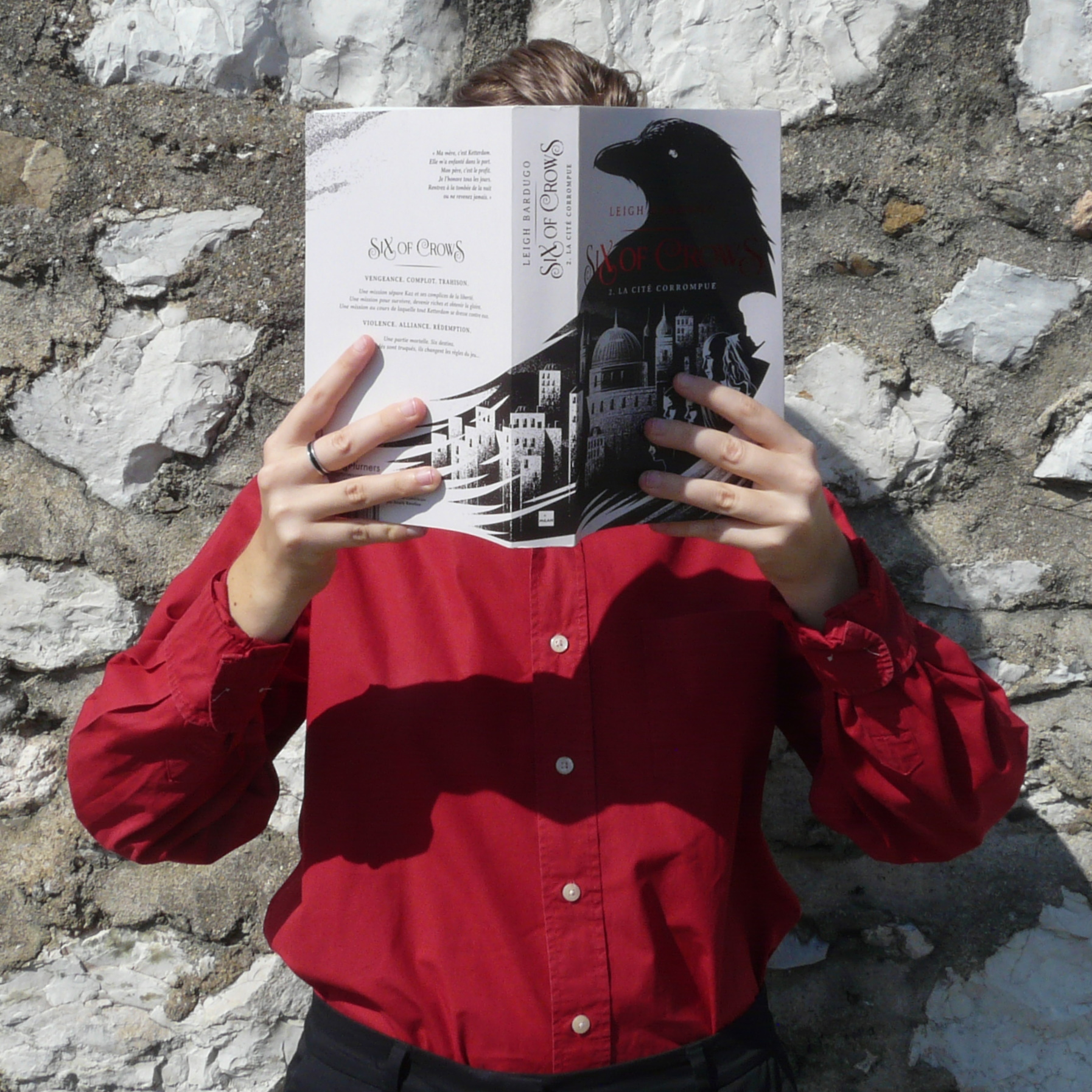 personne en chemise rouge lisant La Cité Corrompue (Six of Crows tome 2) de Leigh Bardugo devant un mur de pierres