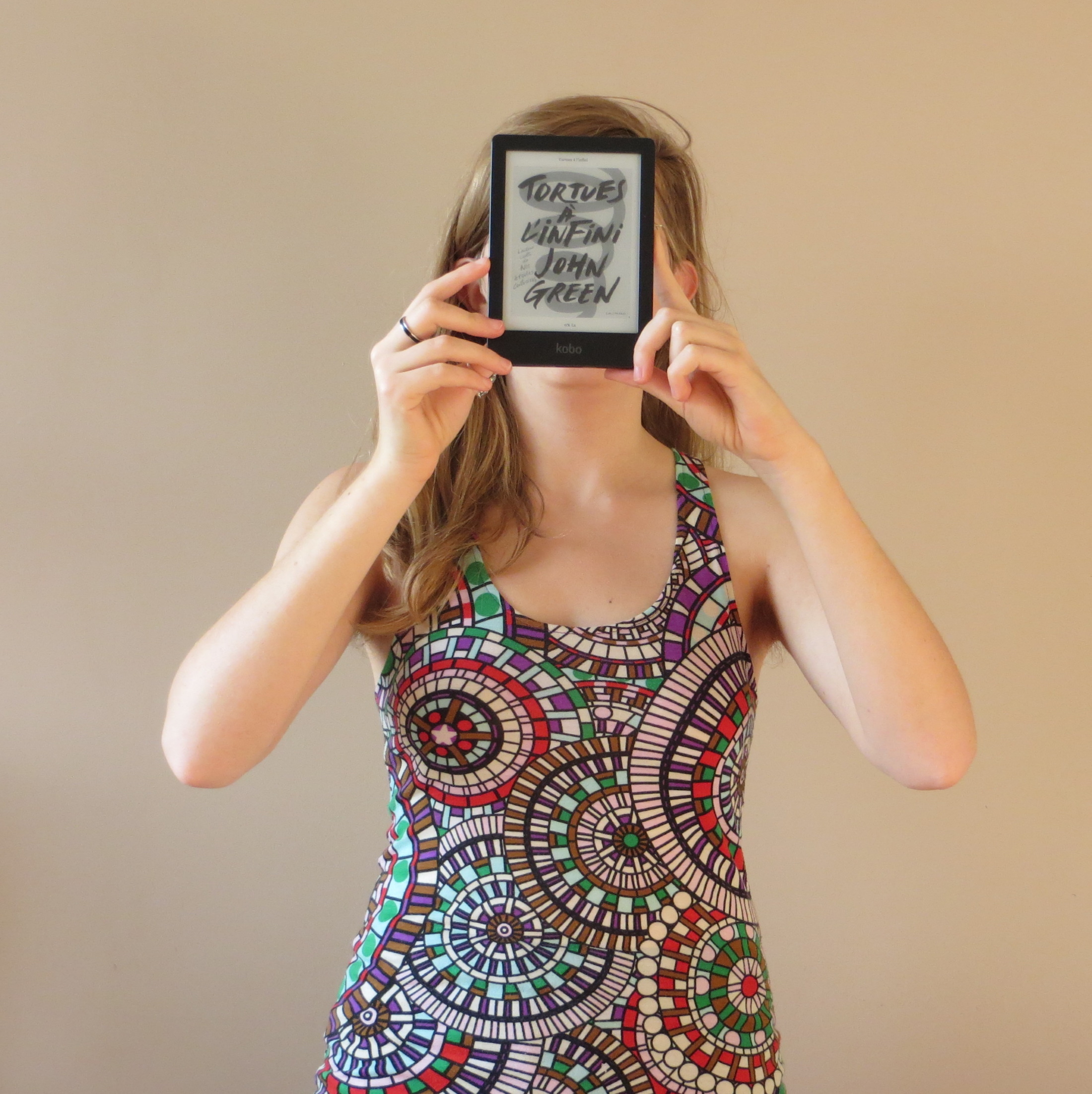 femme en débardeur illusion d'optique brandissant sa liseuse avec la couverture de Tortues à l'Infini de John Green devant un mur beige
