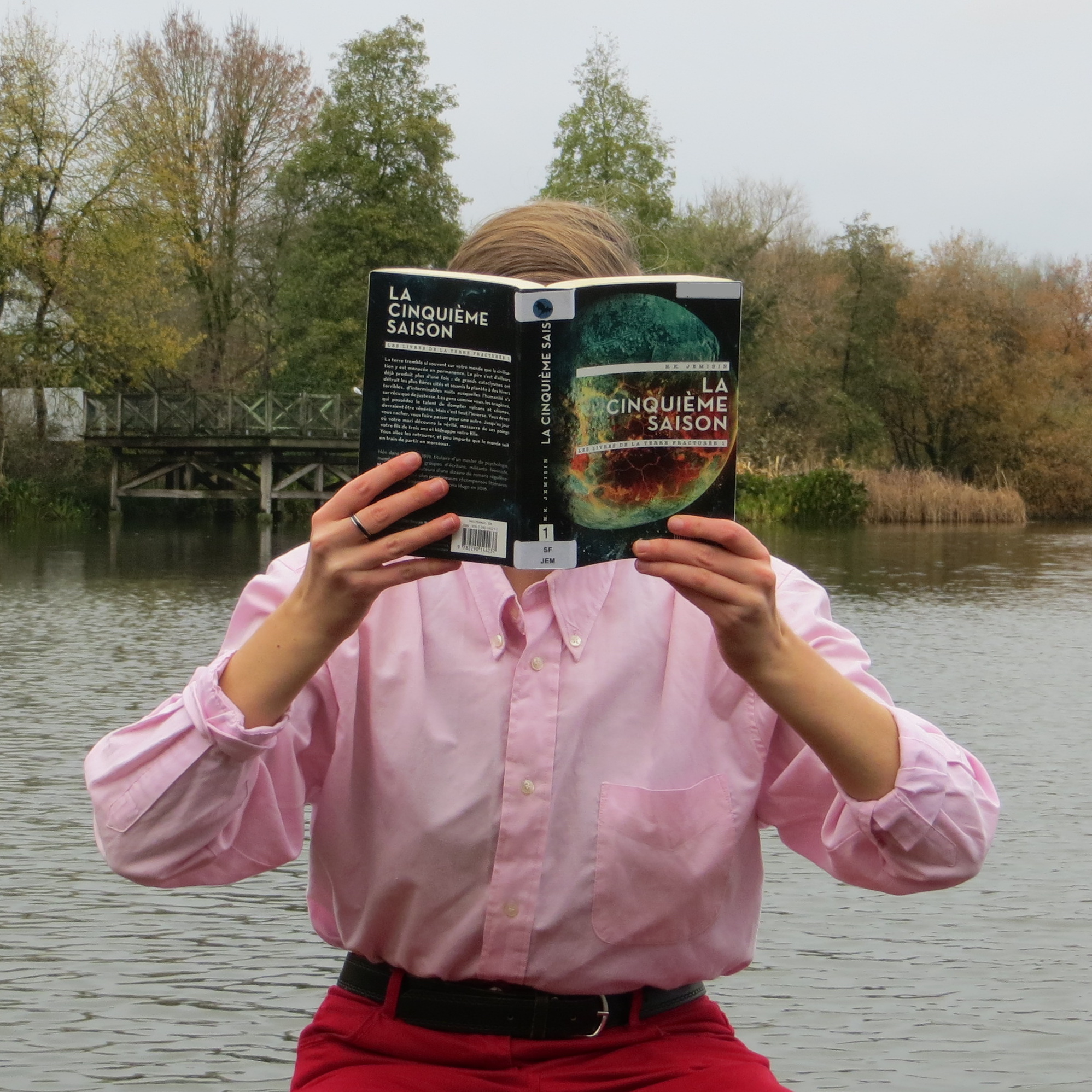 personne en chemise rose lisant Les Livres de la Terre Fracturée tome 1 : La cinquième saison de N.K. Jemisin devant un lac
