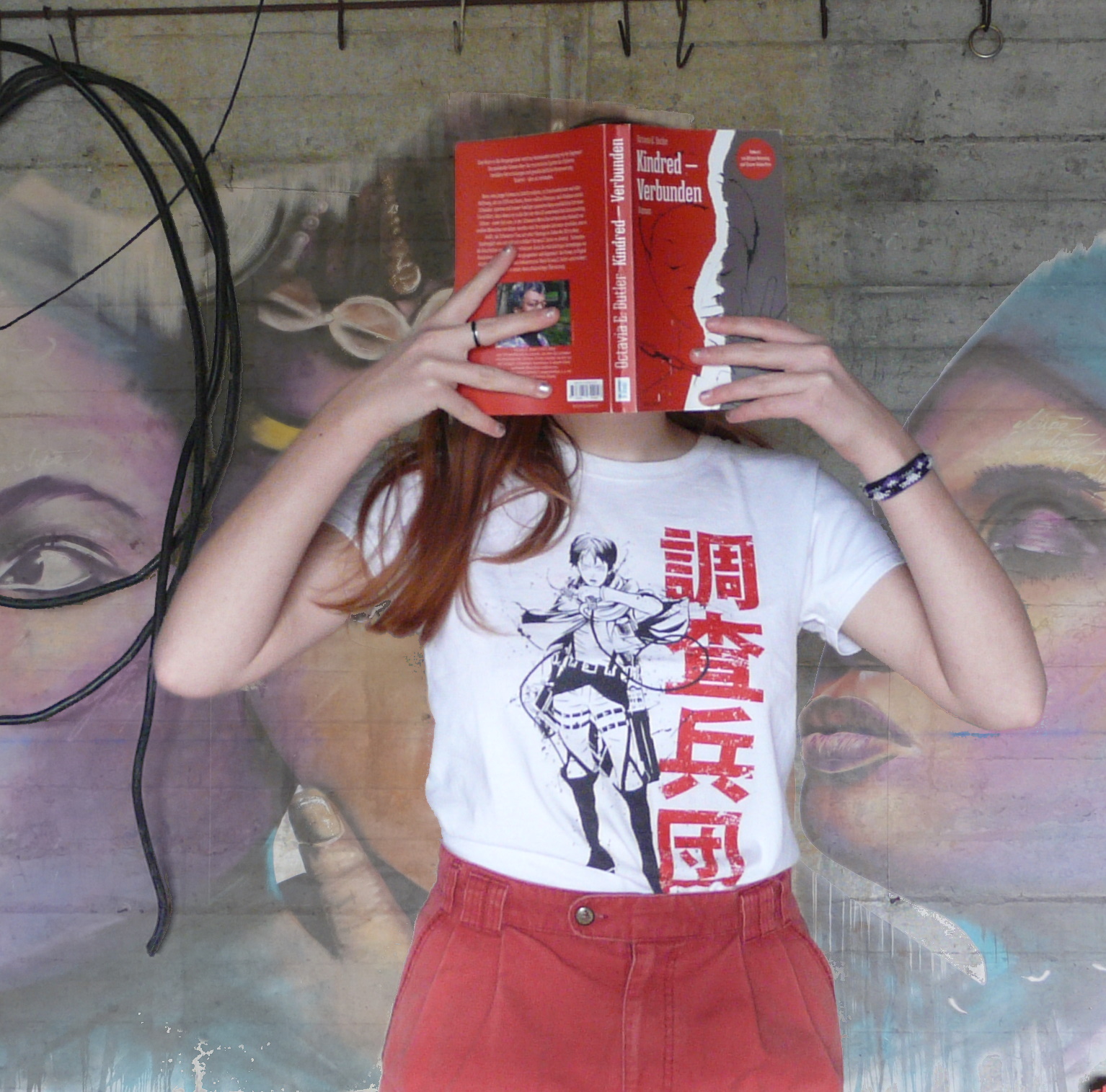 personne en t-shirt attaque des titans et short rouge lisant kindred – Liens de Sang d'Octavia Butler devant un mur tagué