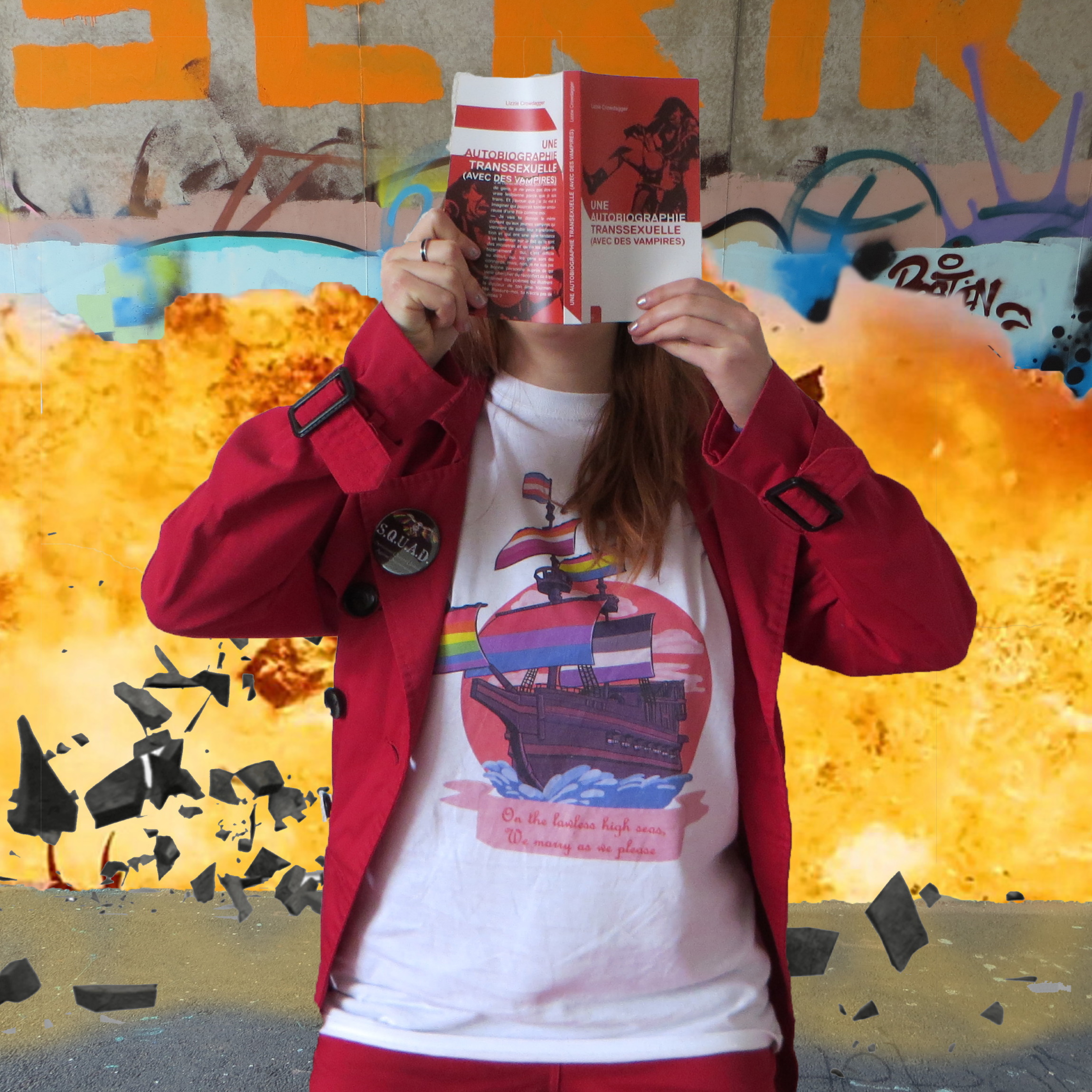 personne en t-shirt pirate queer et parka rouge lisant Une autobiographie transssexuelle devant un mur tagué qui explose