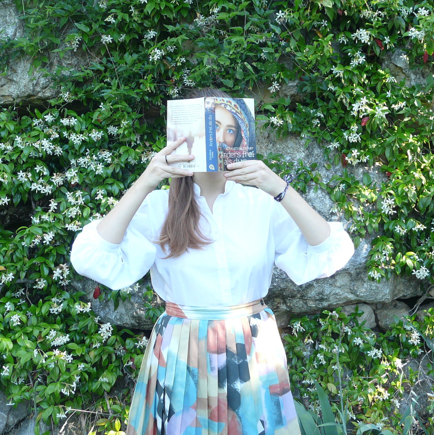 personne en jupe plissée lisant Anders Frei Als Du de Christine Fehèr devant un mur de fleurs blanches