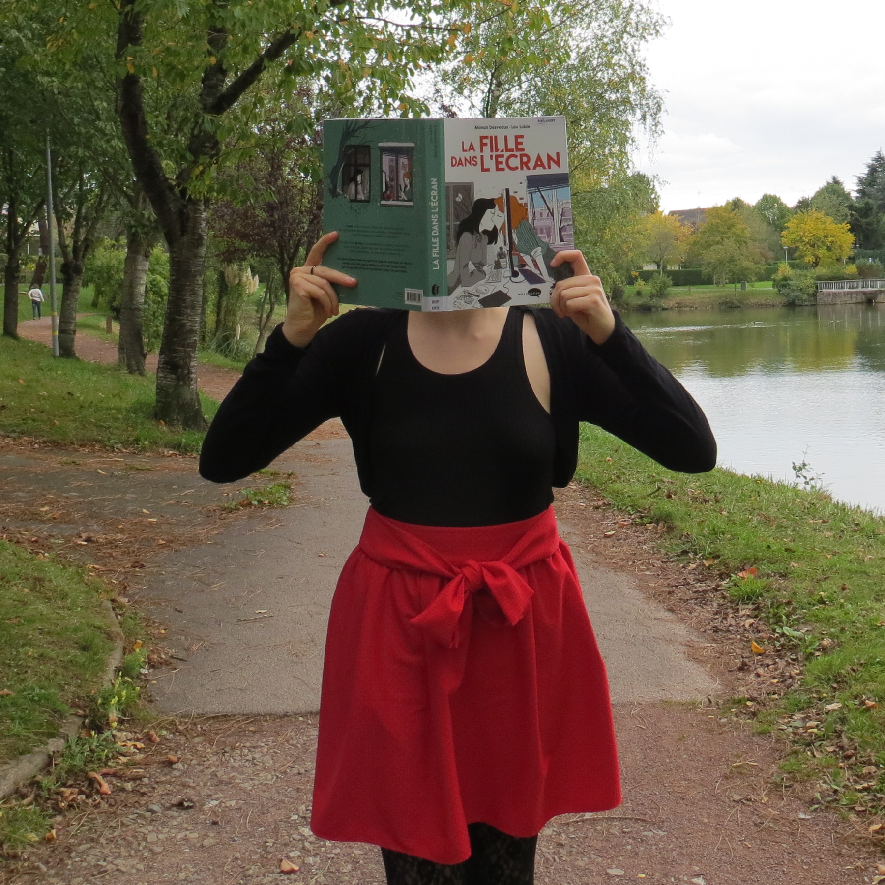 personne en jupe rouge lisant La Fille dans l’Écran de Lou Lubie et Manon Desveaux sur une promenade au bord d'un lac
