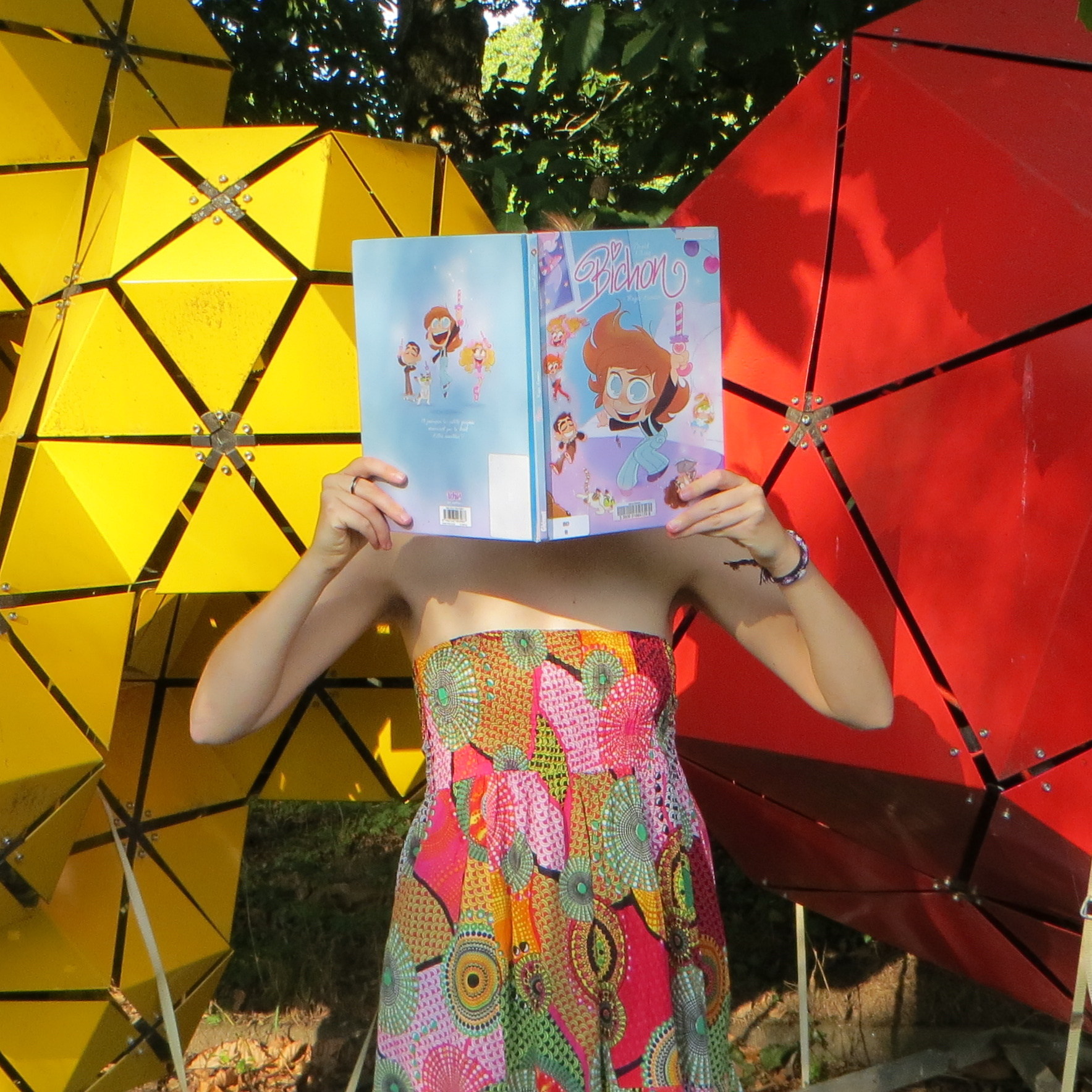 personne en robe multicolore lisant Bichon de David Gilson devant des constructions géométriques colorées
