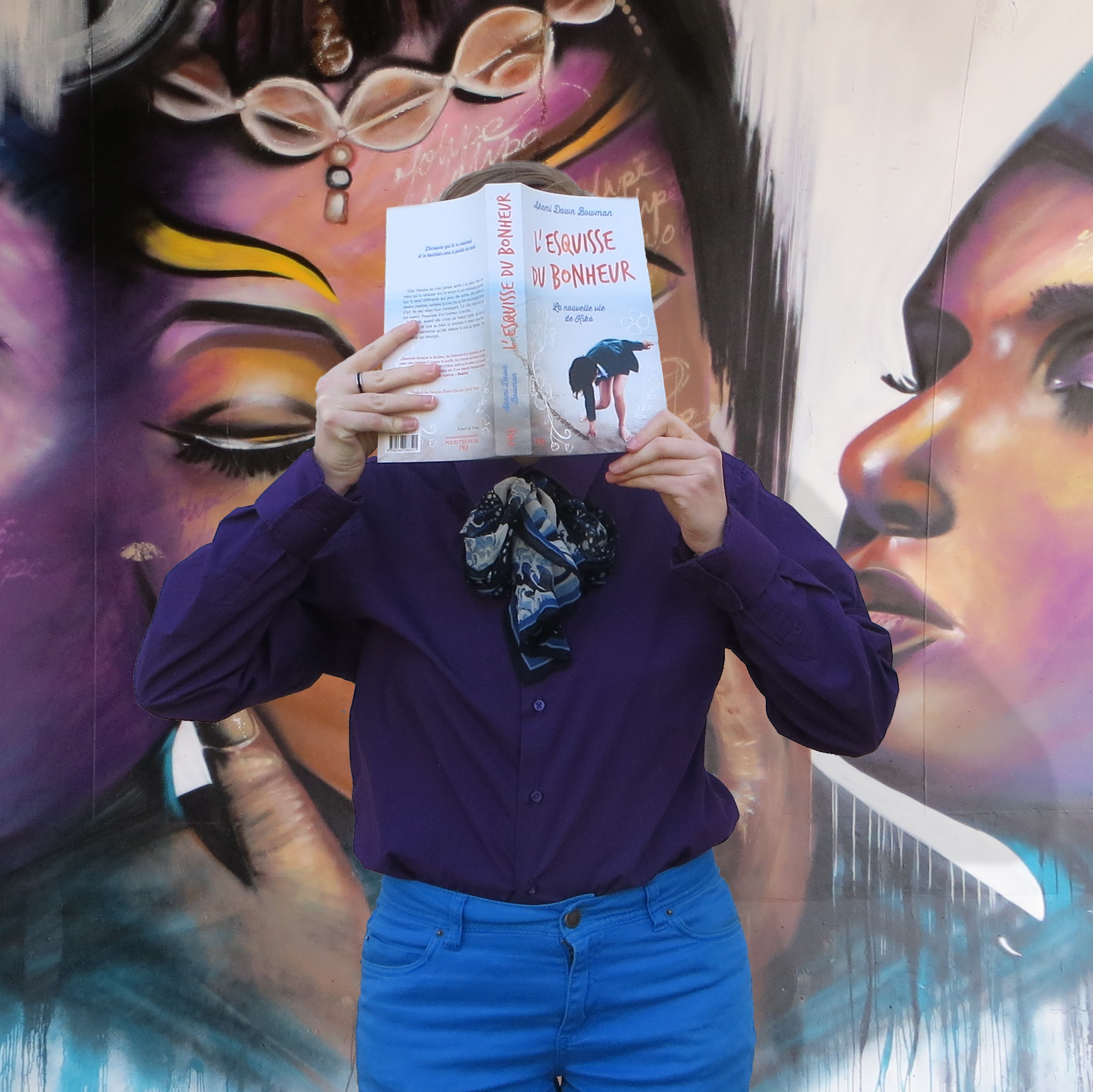 personne en chemise violette et écharpe de La Vague lisant L’esquisse du Bonheur d’Akemi Dawn Bowman devant une fresque murale