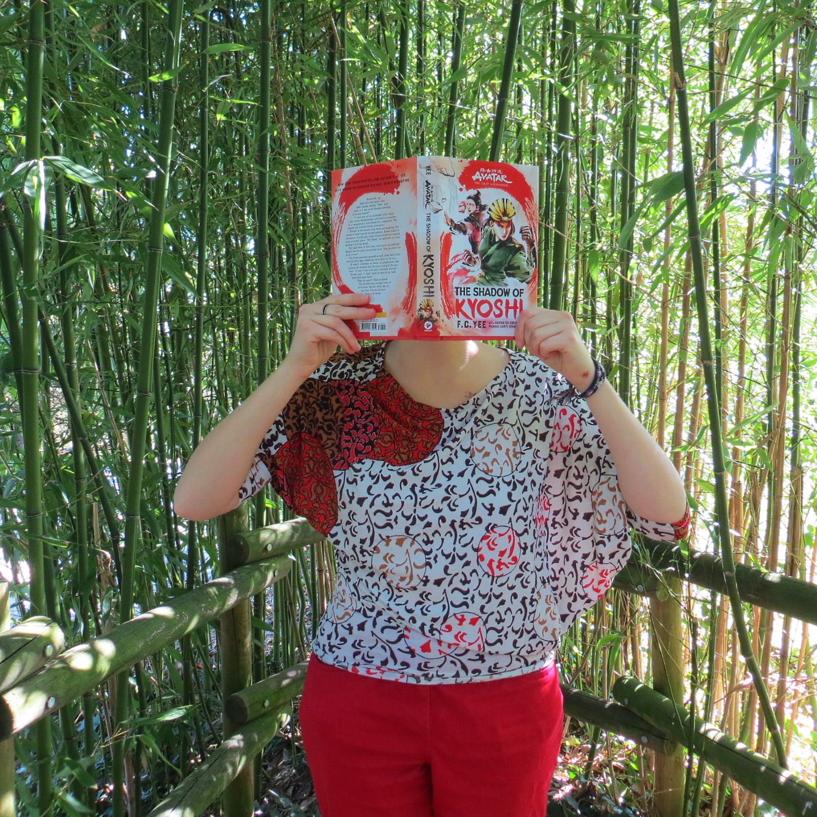personne en chemisier avec un motif de feuilles lisant The Shadow of Kyoshi de F.C. Yee devant une haie de bambous