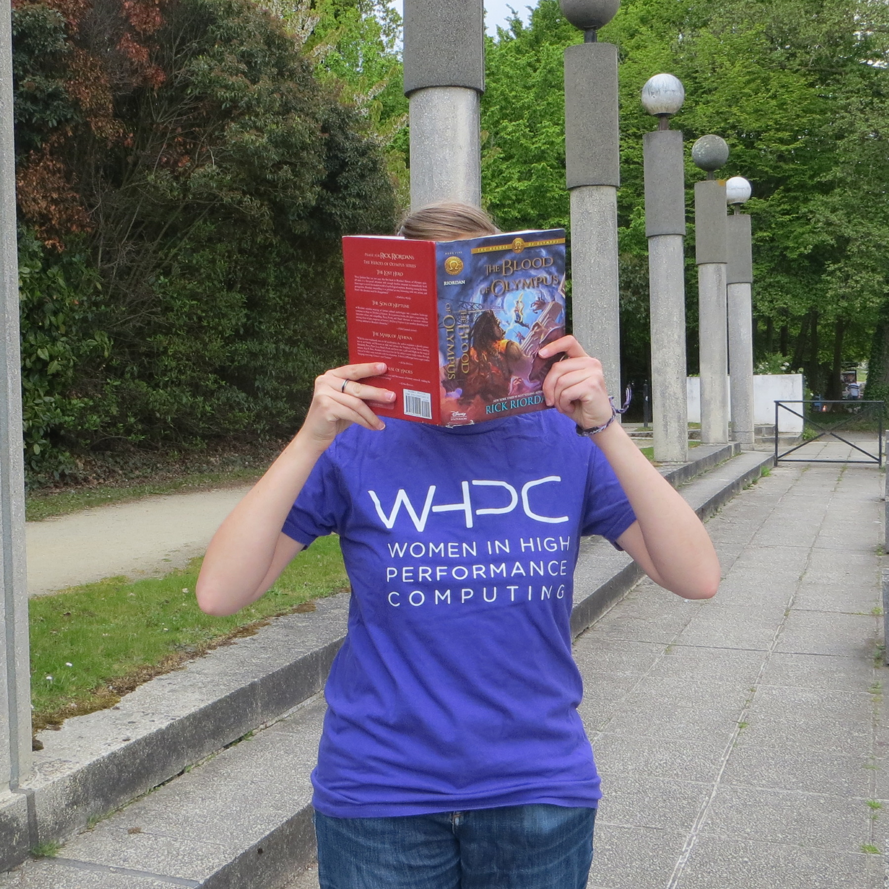 personne en t-shirt violet lisant le tome 5 de Héros de l'Olympe devant une rangée de colonnes