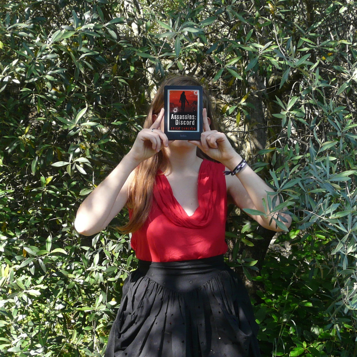 personne en robe noire et rouge tenant une liseuse avec la couverture d'assassins tome 1 : discord d'Erica Cameron, devant un olivier