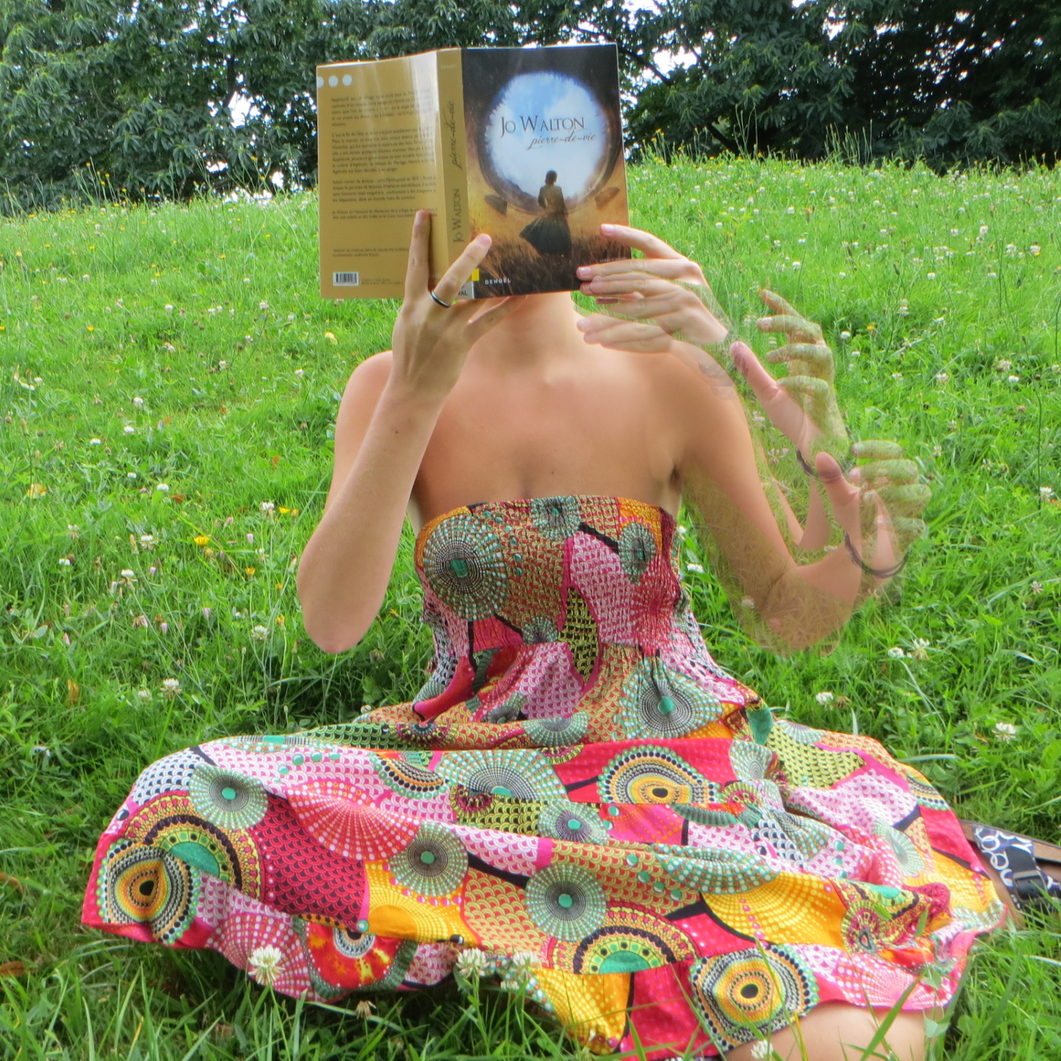 personne en robe colorée lisant Pierre-de-vie de Jo Walton dans l'herbe. On voit en transparence le mouvement de son bras
