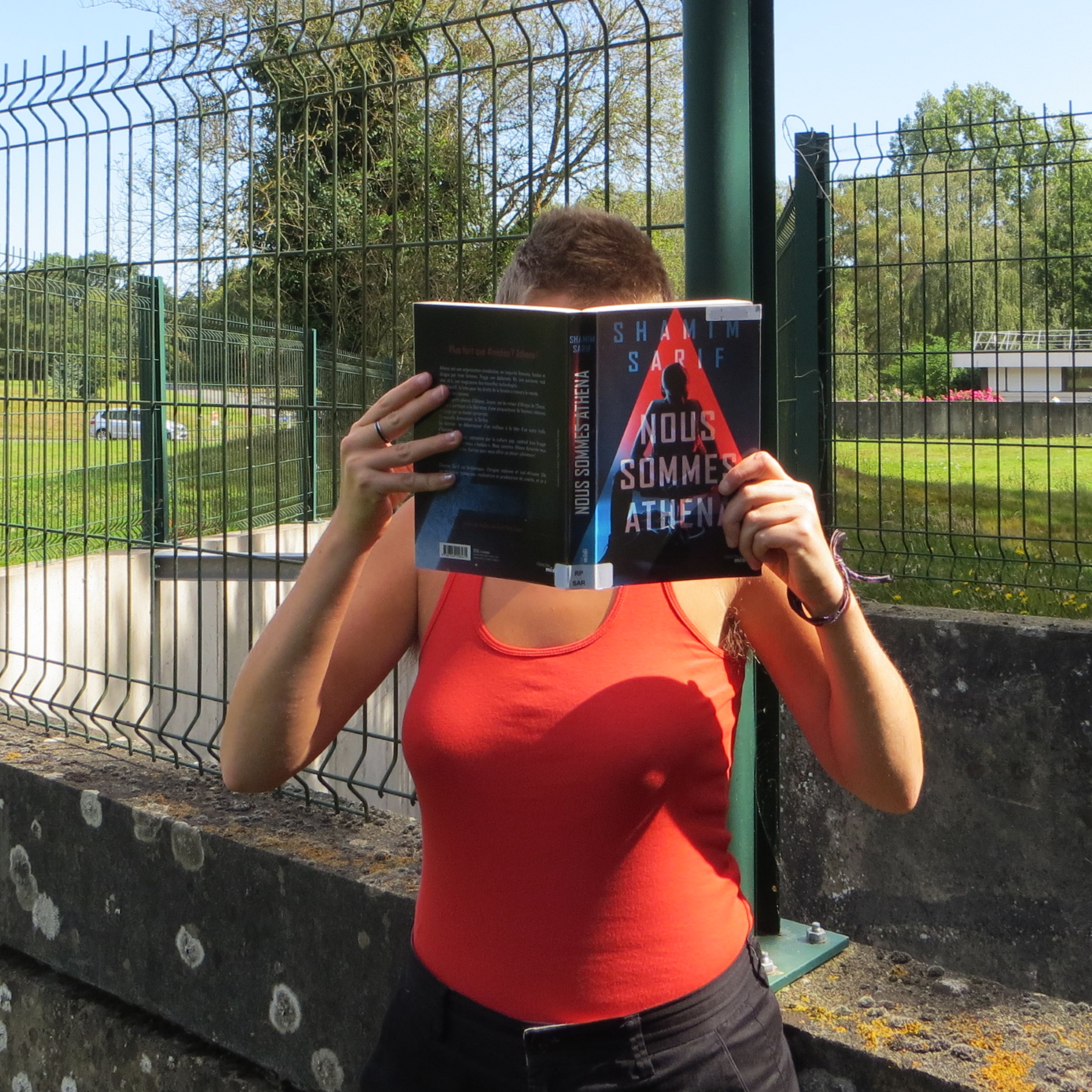 personne en rouge lisant Nous Sommes Athéna de Shamim Sarif devant une grille et des murs