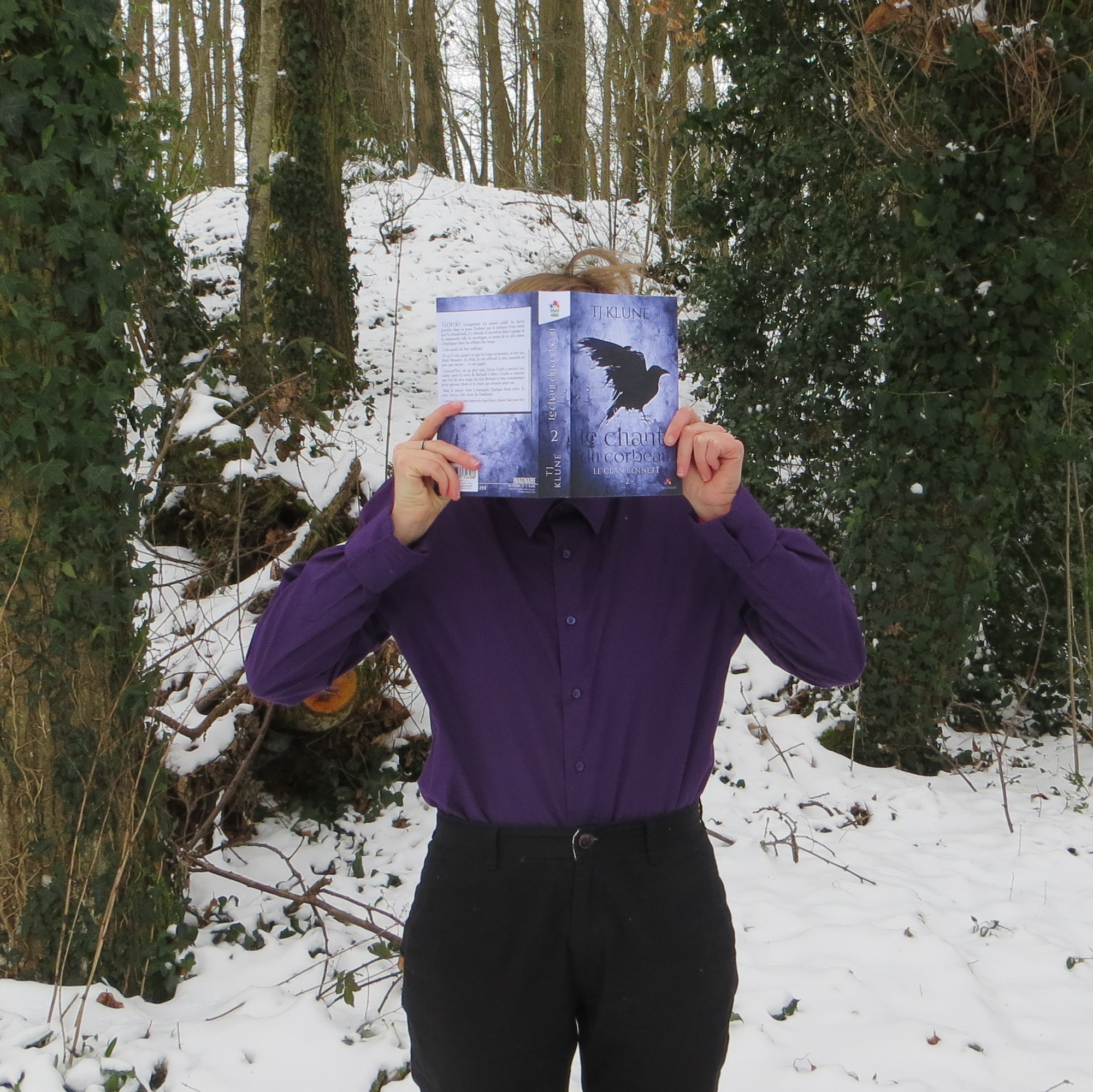 personne en chemise violette lisant Le Chant du Corbeau de T.J. Klune dans une forêt enneigée