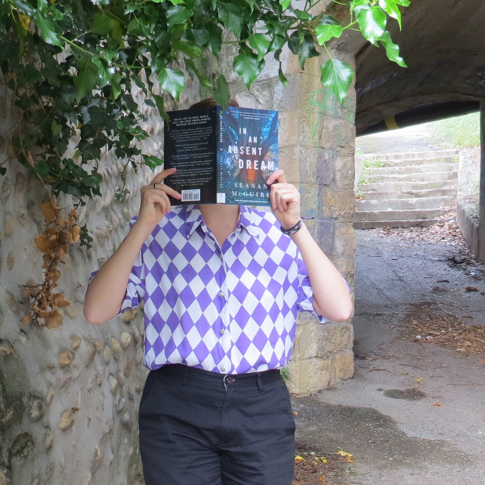 personne en chemise à carreaux violets lisant Wayward Children Tome 4 : In an Absent Dream de Seanan McGuire devant un petit tunnel