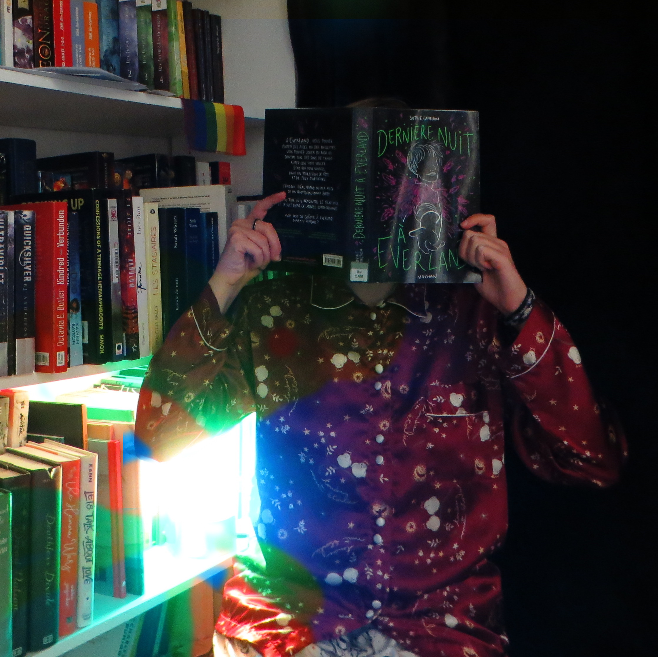 personne en pyjama lisant Dernière nuit à Everland de Sophie Cameron devant des étagères de livres avec des lumières multicolores