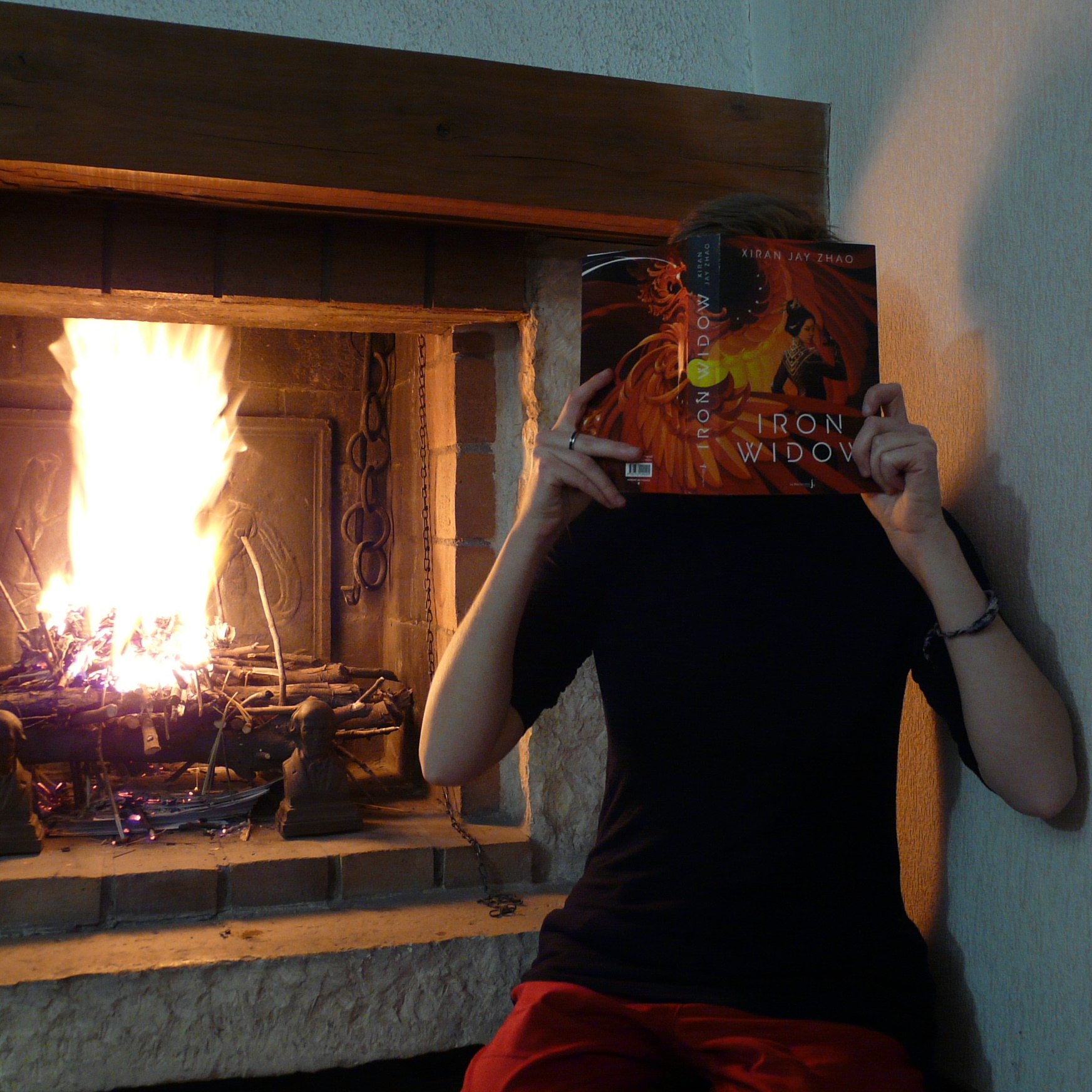 personne en rouge et noir lisant Iron Widow de Xiran Jay Zhao devant un feu de cheminée