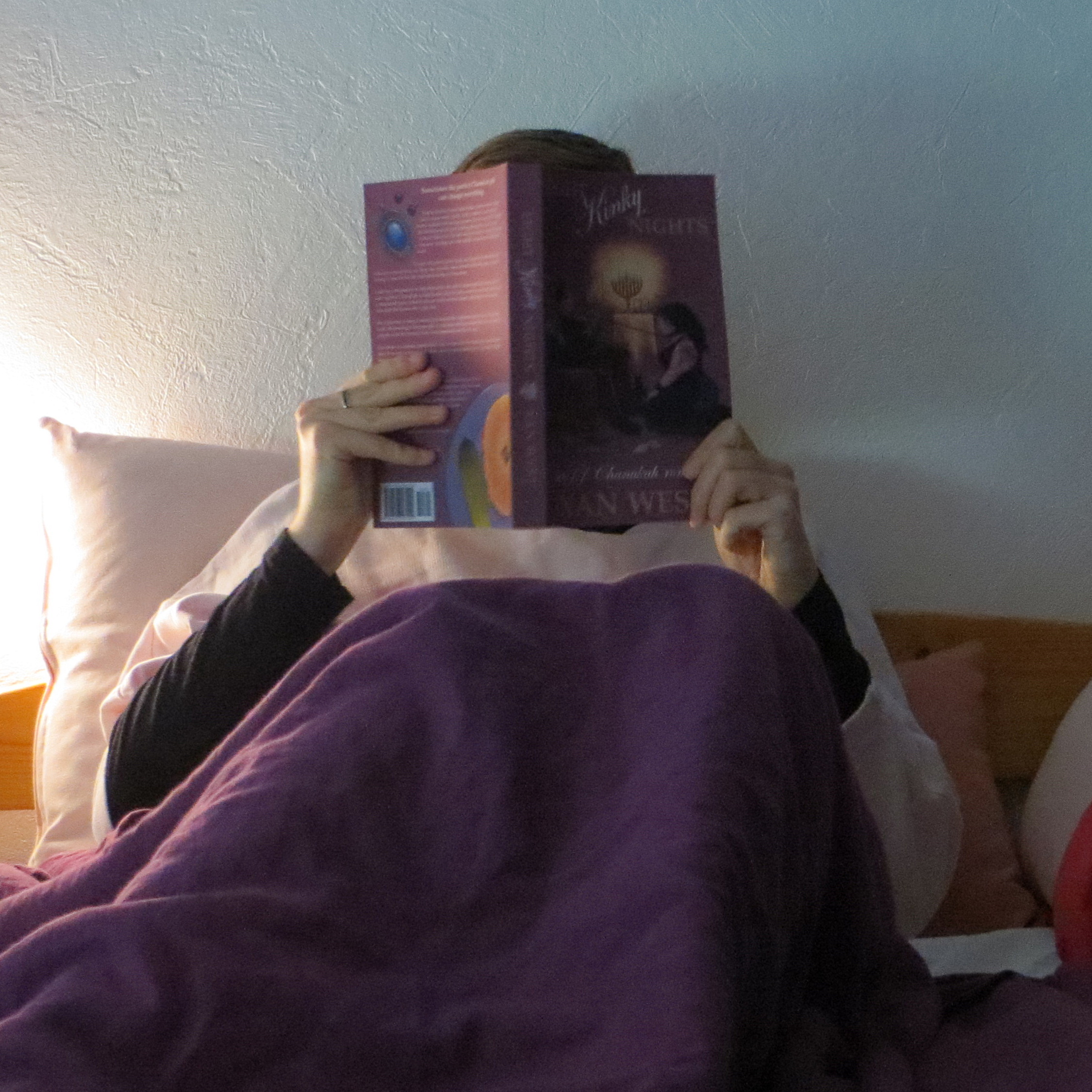 personne en pull doux rose lisant Eight Kinky Nights de Xan West dans son lit, avec une couverture violette