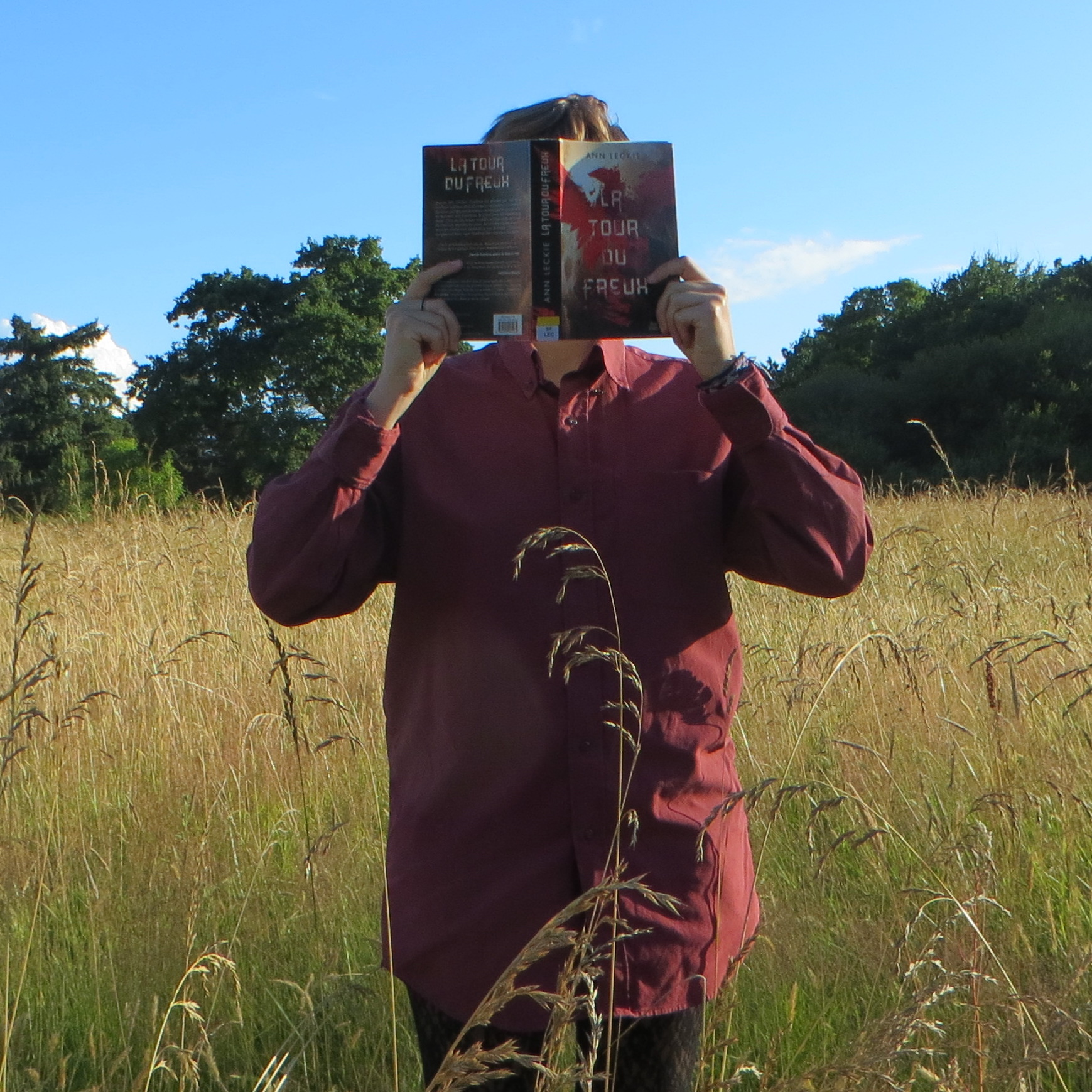 personne en chemise rouge lisant La Tour du Freux de Ann Leckie devant un champ de blé