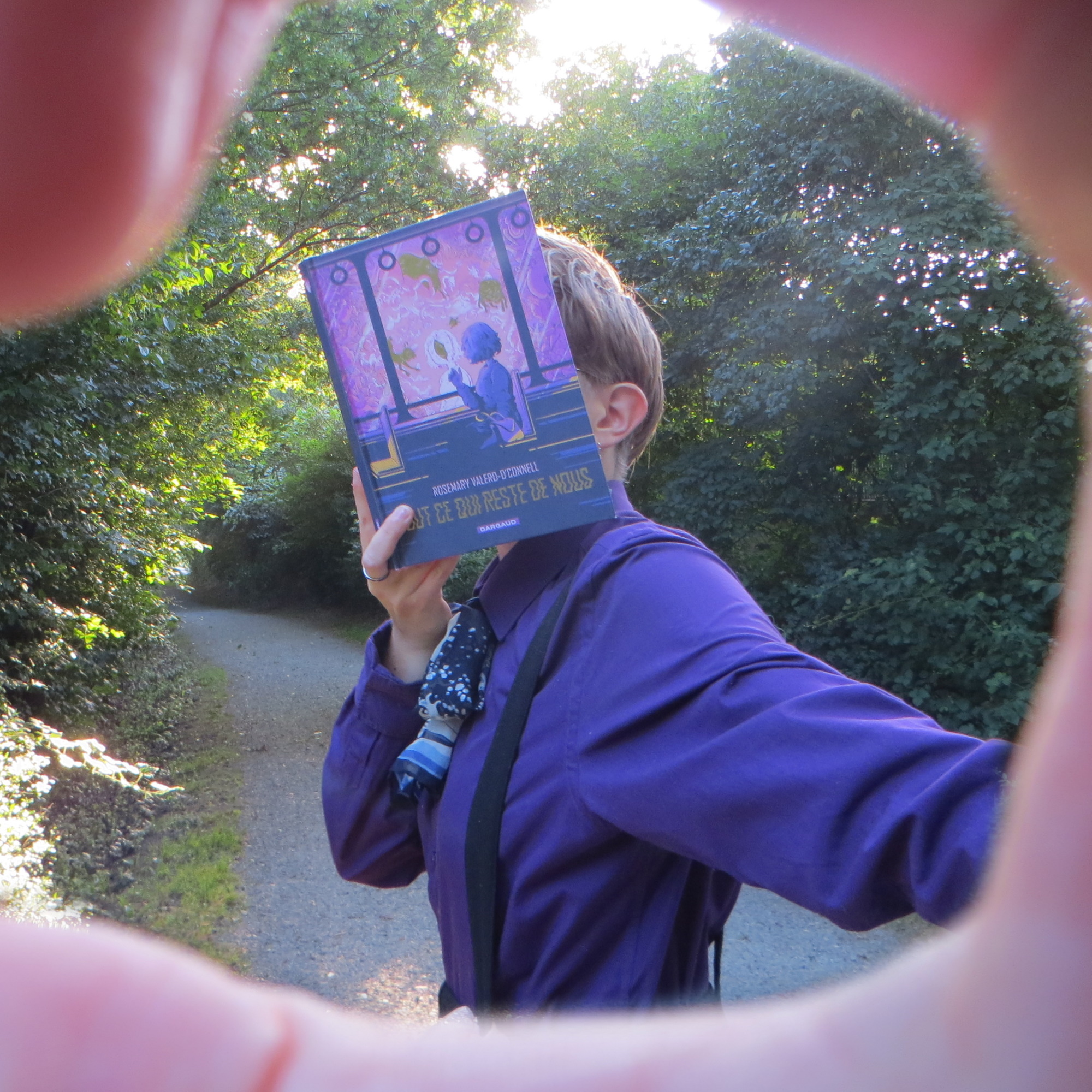à travers un cadre entouré d'une main, on voit une personne en chemise violette lisant Tout ce qui Reste de Nous de Rosemary Valero O’Connell devant un chemin entouré d'arbres au soleil couchant