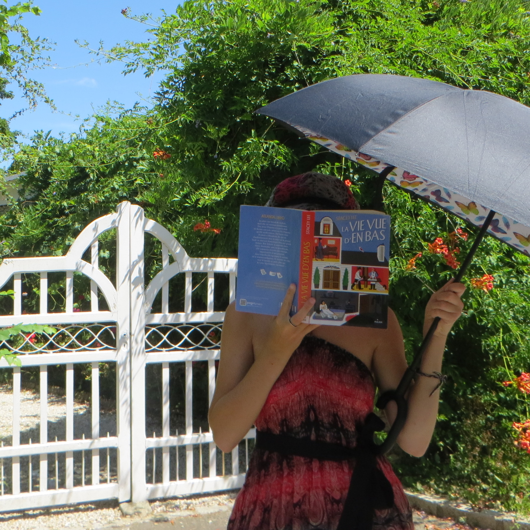 personne en robe rouge et noire, ruban et chapeau, tenant un parapluie, lisant La Vie Vue d’en Bas de Stacey Lee devant un portail entouré de plantes