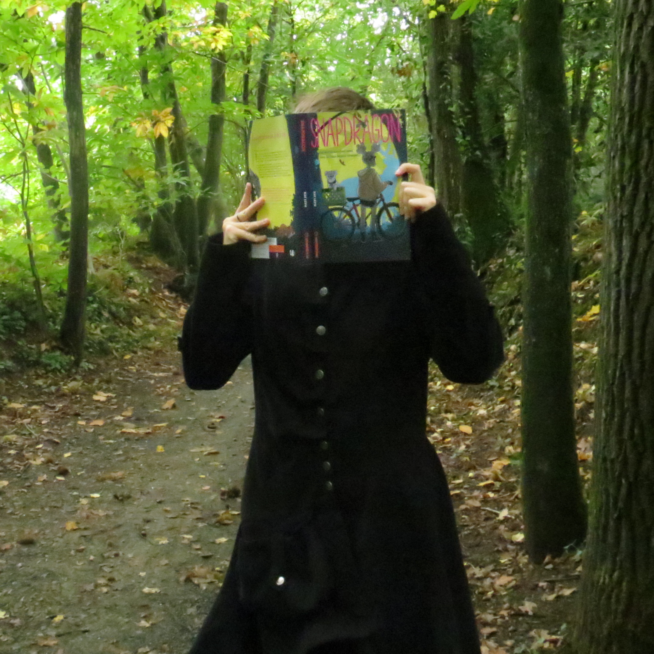 personne en long manteau noir lisant Snapdragon de Kat Leyh dans une forêt pleine de feuilles mortes
