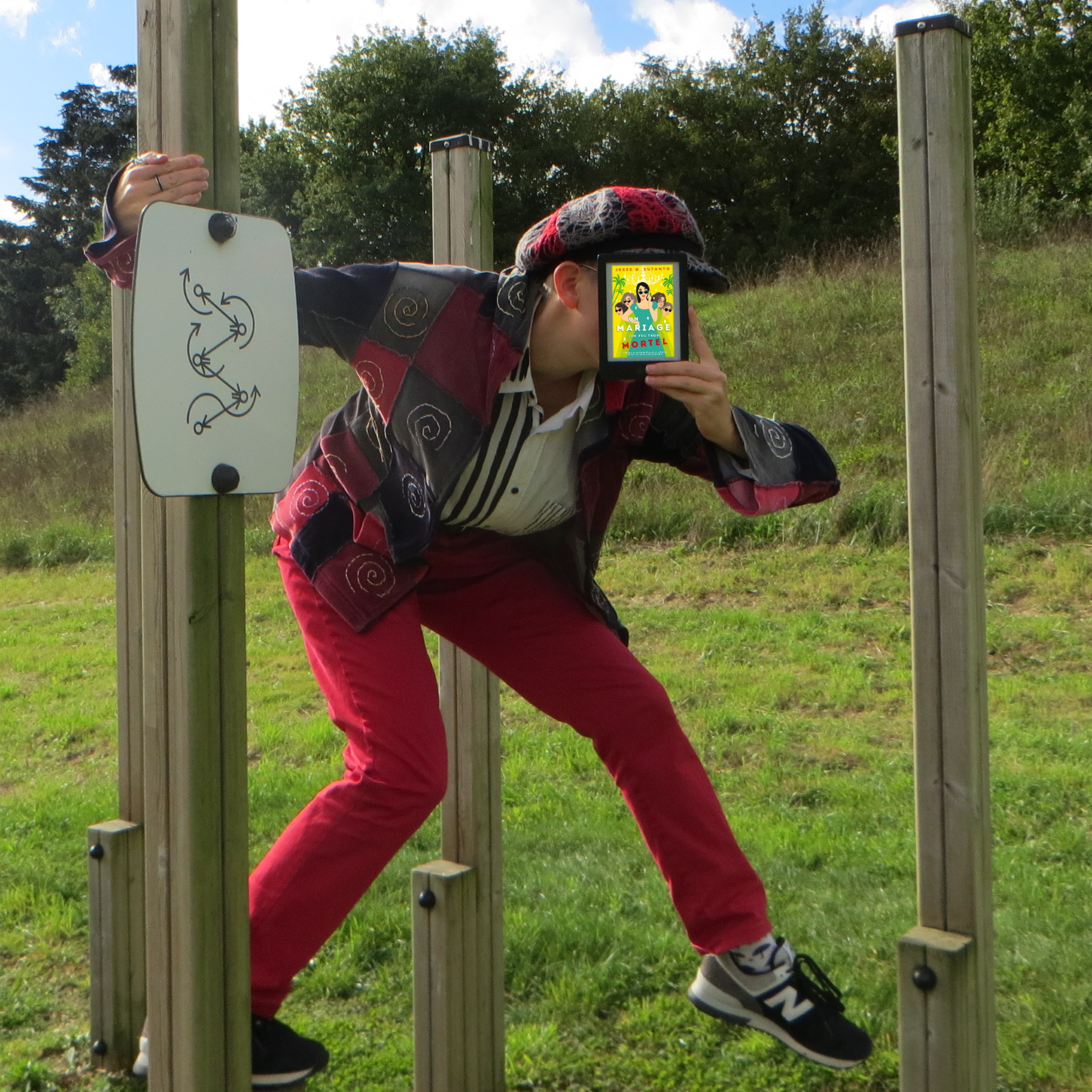 personne en tenue rouge asymétrique progressant sur un parcours d'équilibre en bois, tenant une liseuse avec la couverture de Un Mariage (un peu) trop Mortel de Jesse Q. Sutanto