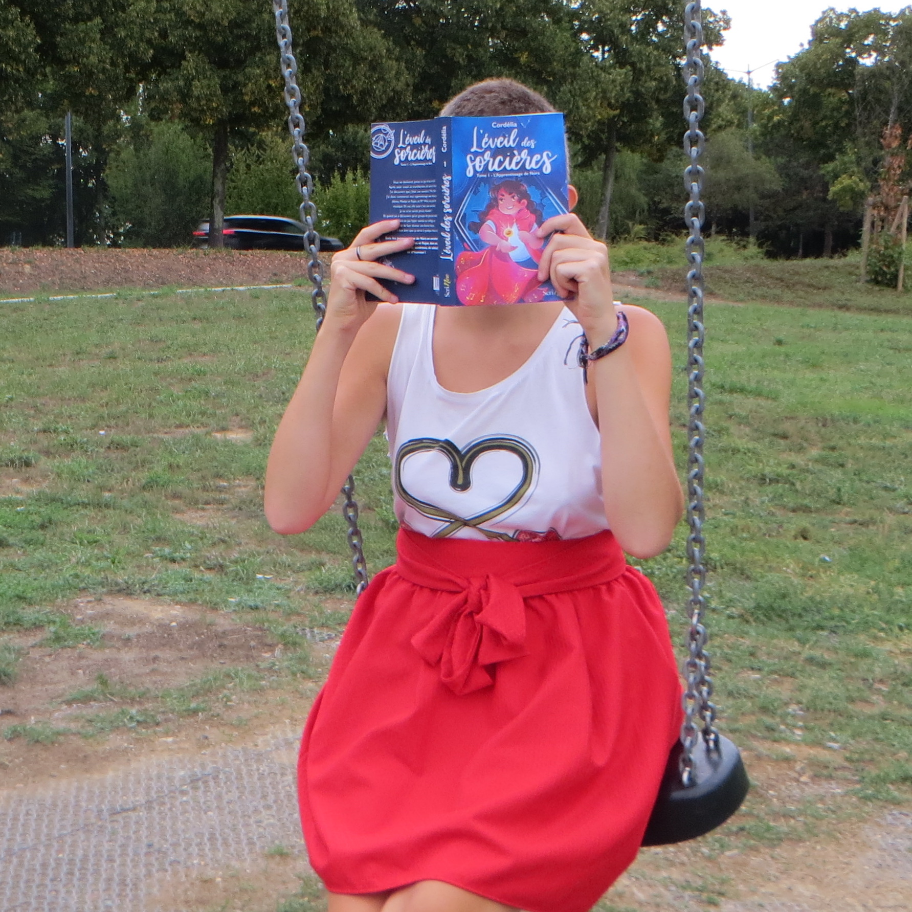 personne en t-shirt cerise-cœur et jupe rouge lisant L'Éveil des Sorcières 1 : L'apprentissage de Nora de Cordélia sur une balançoire