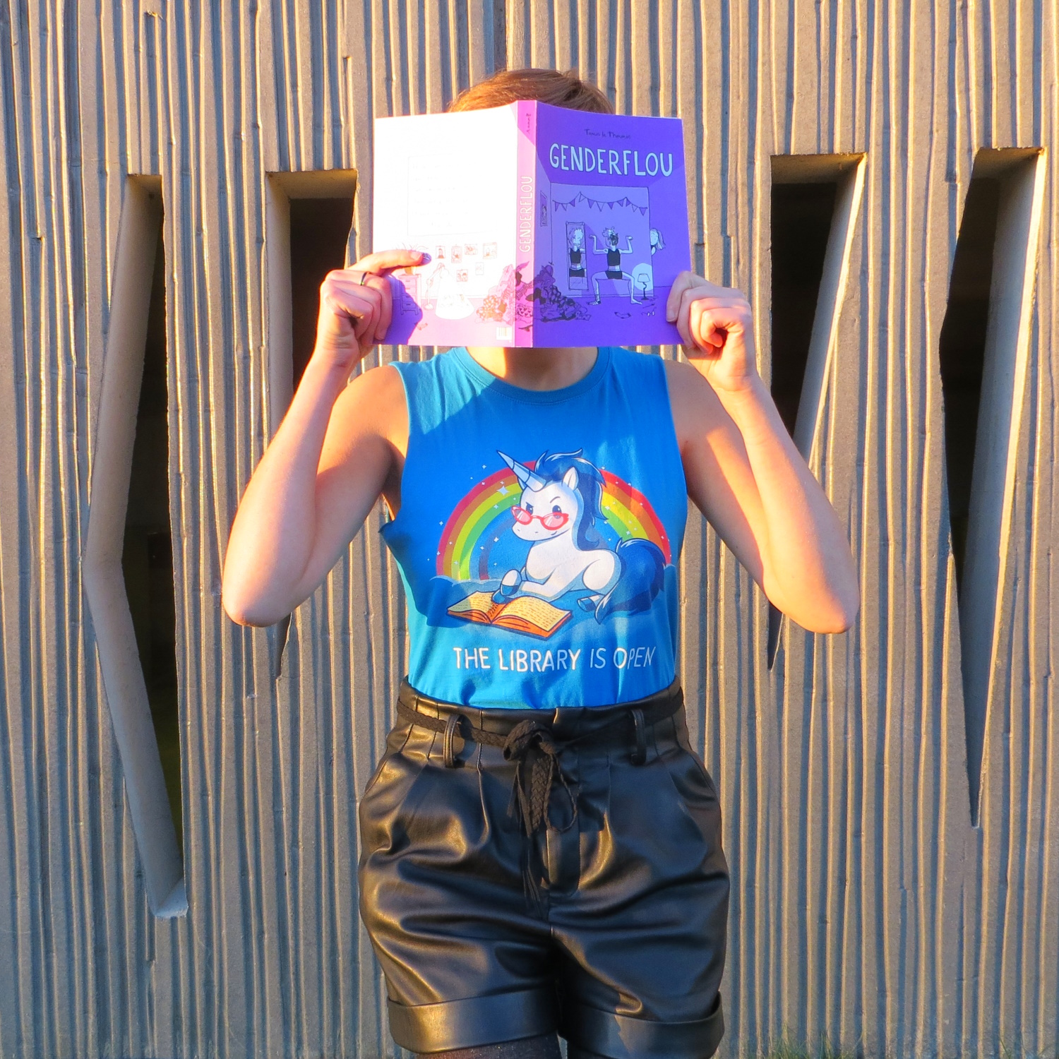 personne en t-shirt licorne lisant Genderflou de Tamos le Thermos devant un mur troué