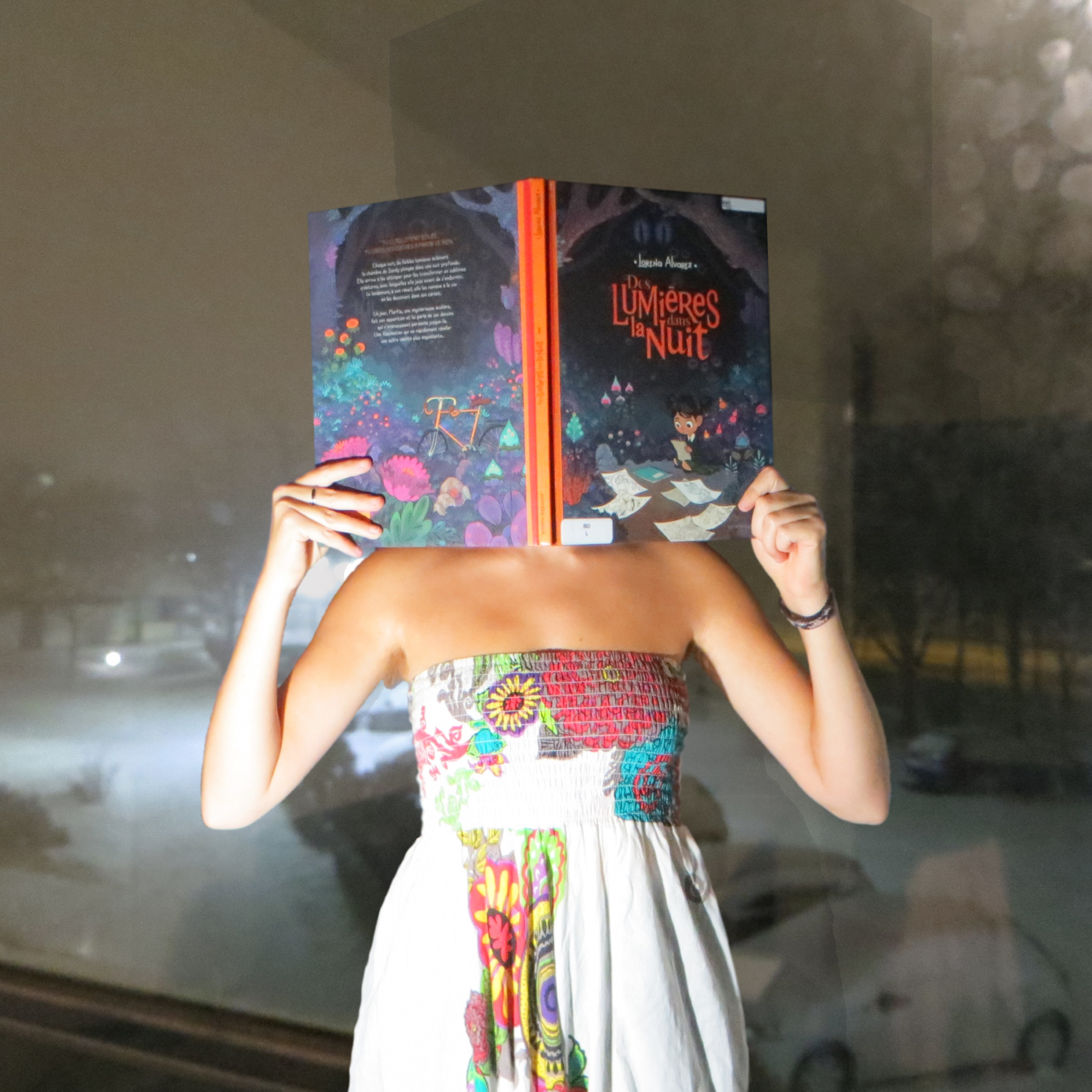 personne en sarouel blanc aux motifs colorés et abstraits lisant Des Lumières dans la nuit de Lorena Álvarez devant une fenêtre donnant sur un parking enneigé