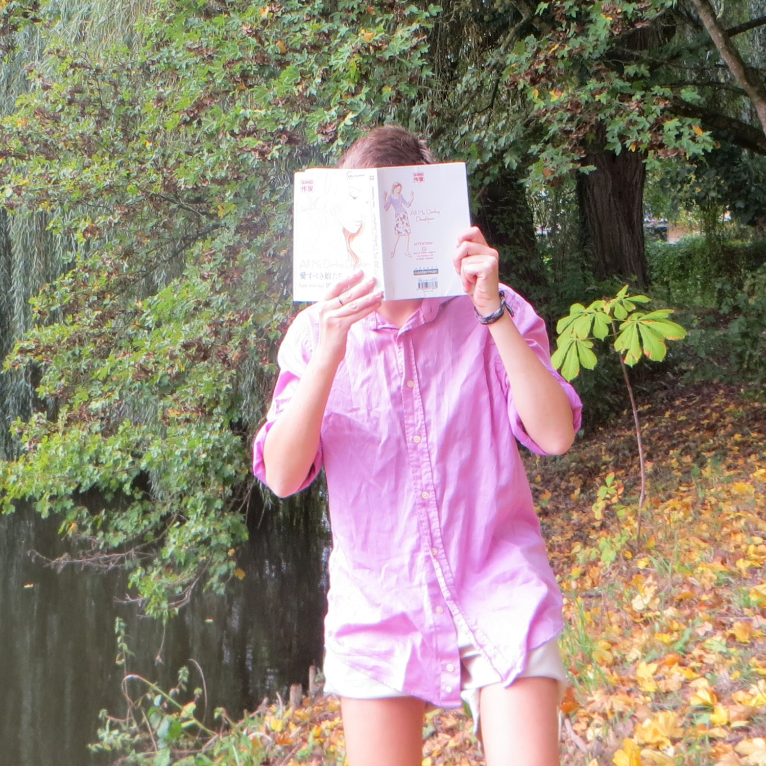 personne en chemise rose et short lisant All my Darlings Daughters de Fumi Yoshinaga devant une rivière et berge de feuilles mortes