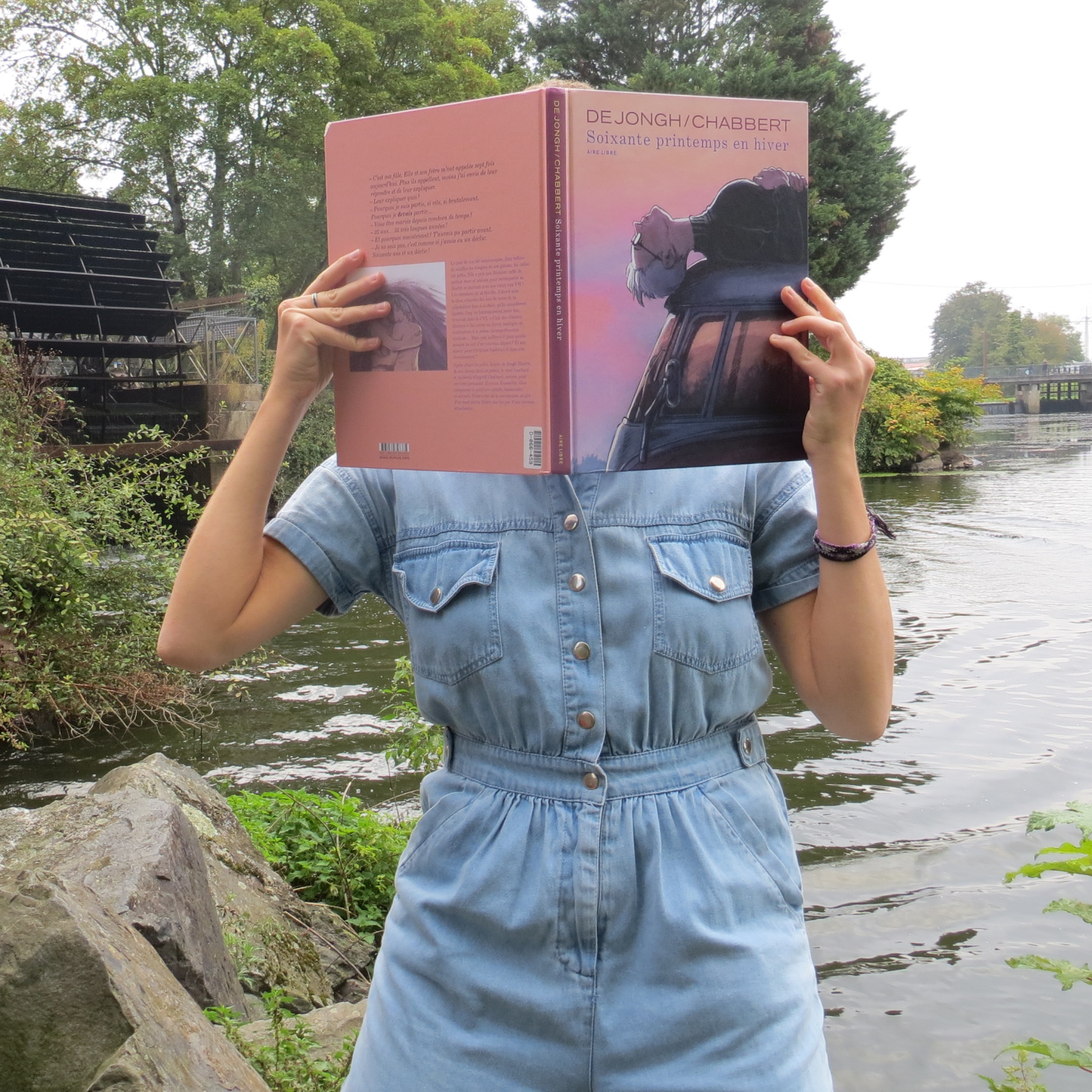 personne en combi en jean lisant Soixante Printemps en Hiver d’Ingrid Chabbert & de Jongh devant un moulin à eau
