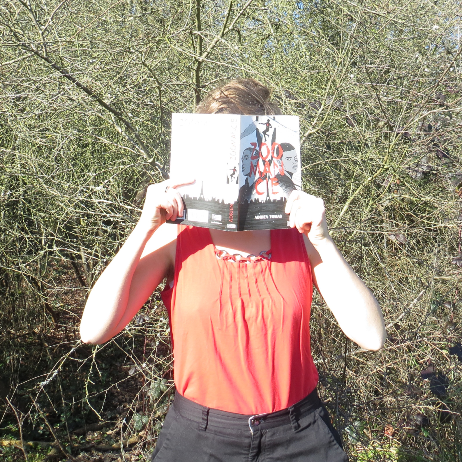 personne en débardeur rouge lisant Zoomancie de Adrien Tomas devant un buisson sans feuilles