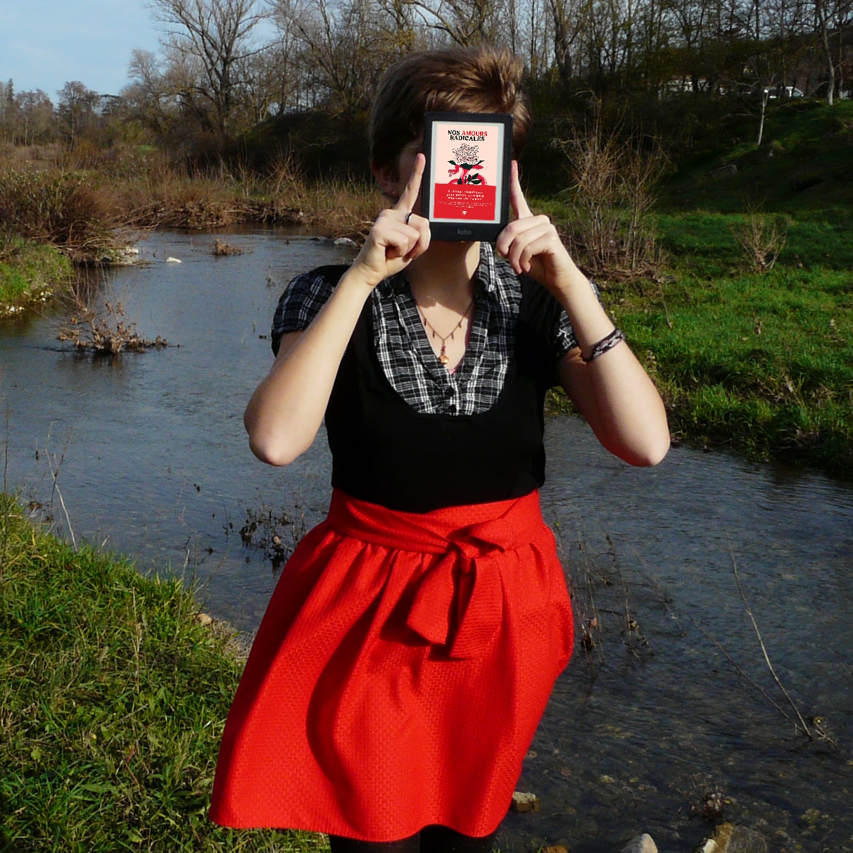 personne en jupe rouge et haut noir lisant Nos Amours Radicales devant une rivière