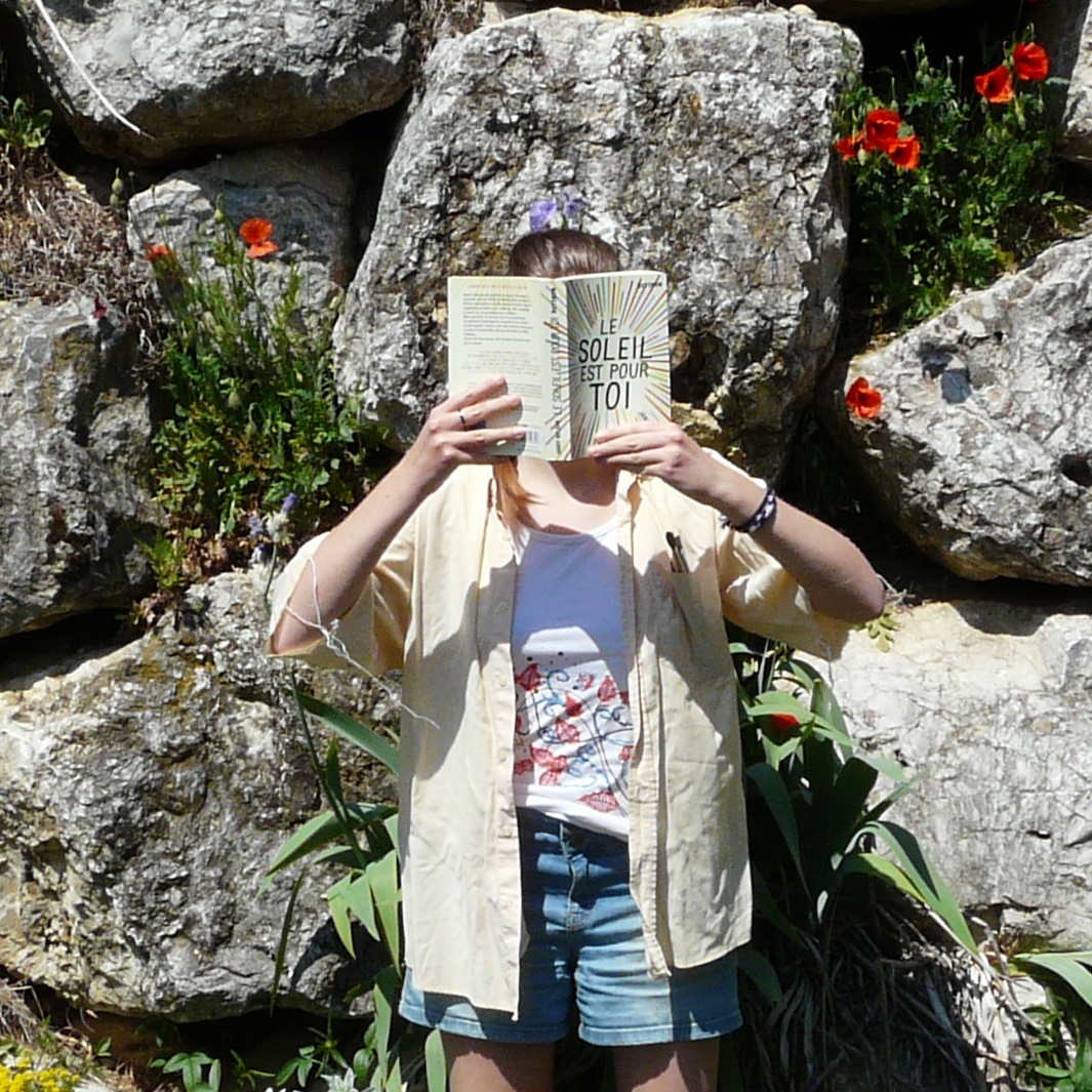 personne en chemise jaune pale ouverte et short lisant Le soleil est pour toi de Jandy Nelson devant des rochers pleins de fleurs