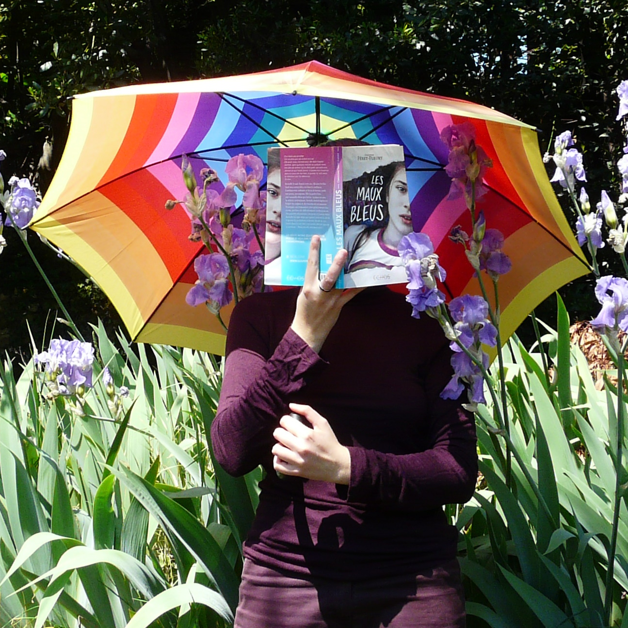 personne en violet lisant Les Maux Bleus de Christine Féret-Fleury en tenant un parapluie arc-en-ciel devant des iris violets