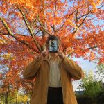 La Fille de Braise et de Ronces de Rae Carson personne en manteau jaune lisant La Fille de Braise et de Ronces de Rae Carson devant un arbre aux couleurs d'automne