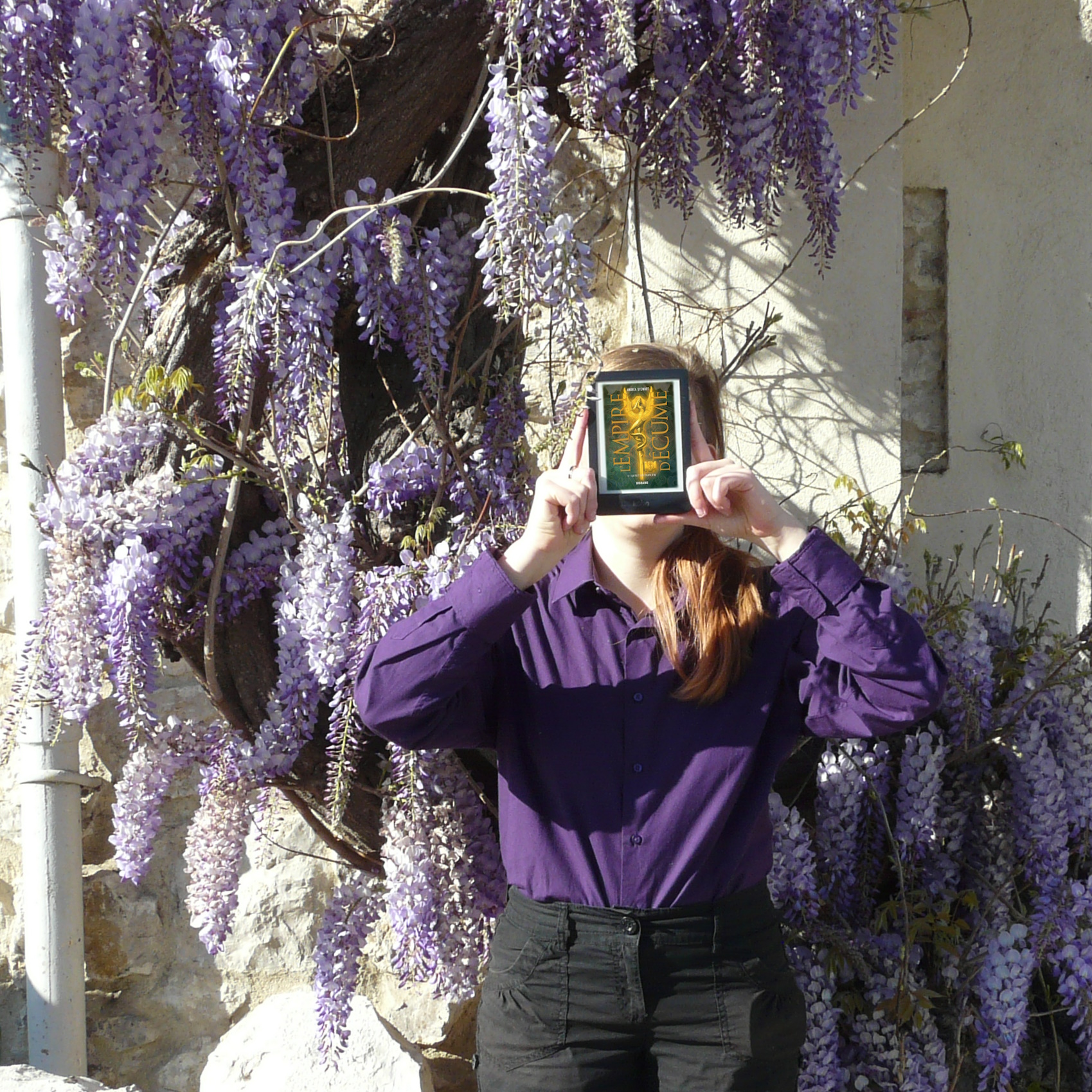 personne en chemise violette lisant L’Empire d’Écume tome 1 : La Fille aux Éclats d’Os de Andrea Stewart devant une plante grimpante aux fleurs violettes