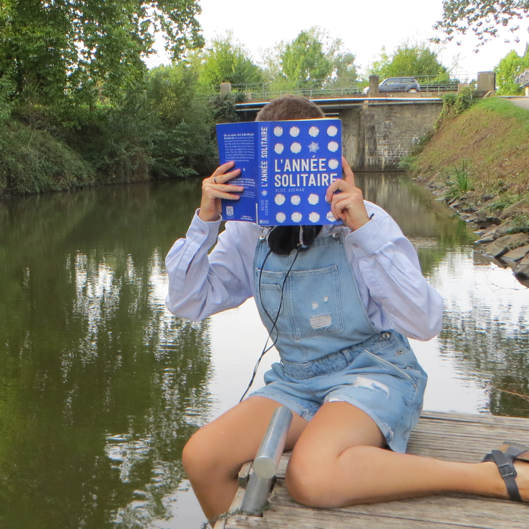 personne en salopette et chemise lisant L’année Solitaire d’Alice Oseman sur un ponton devant une rivière