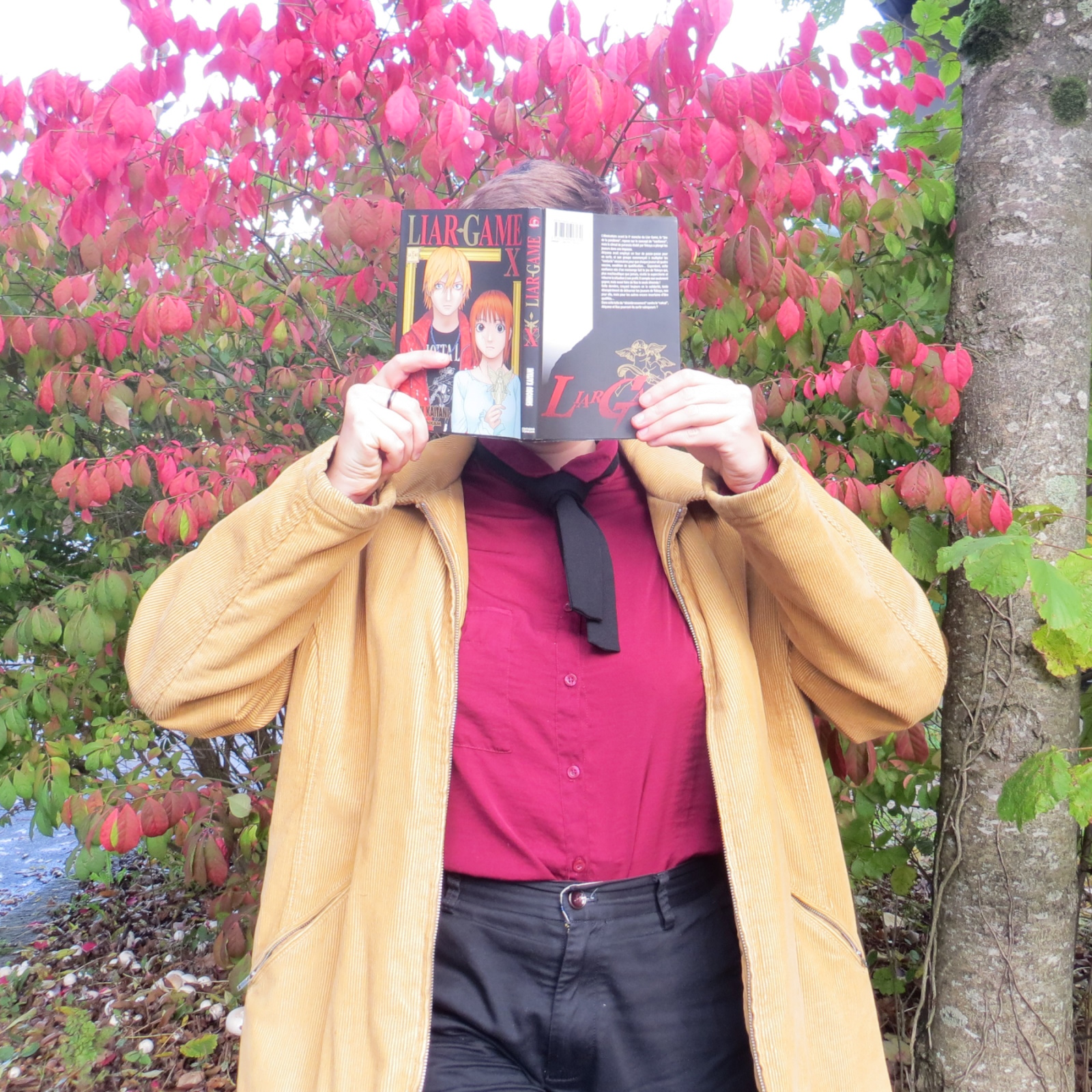 personne en chemise rouge et veste jaune lisant Liar Game de Shinobu Kaitani devant un buisson rouge