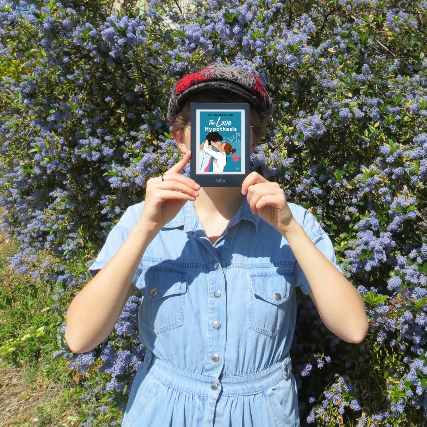 personne en combi de jean lisant The Love Hypothesis de Ali Hazelwood devant un buisson de fleurs bleues