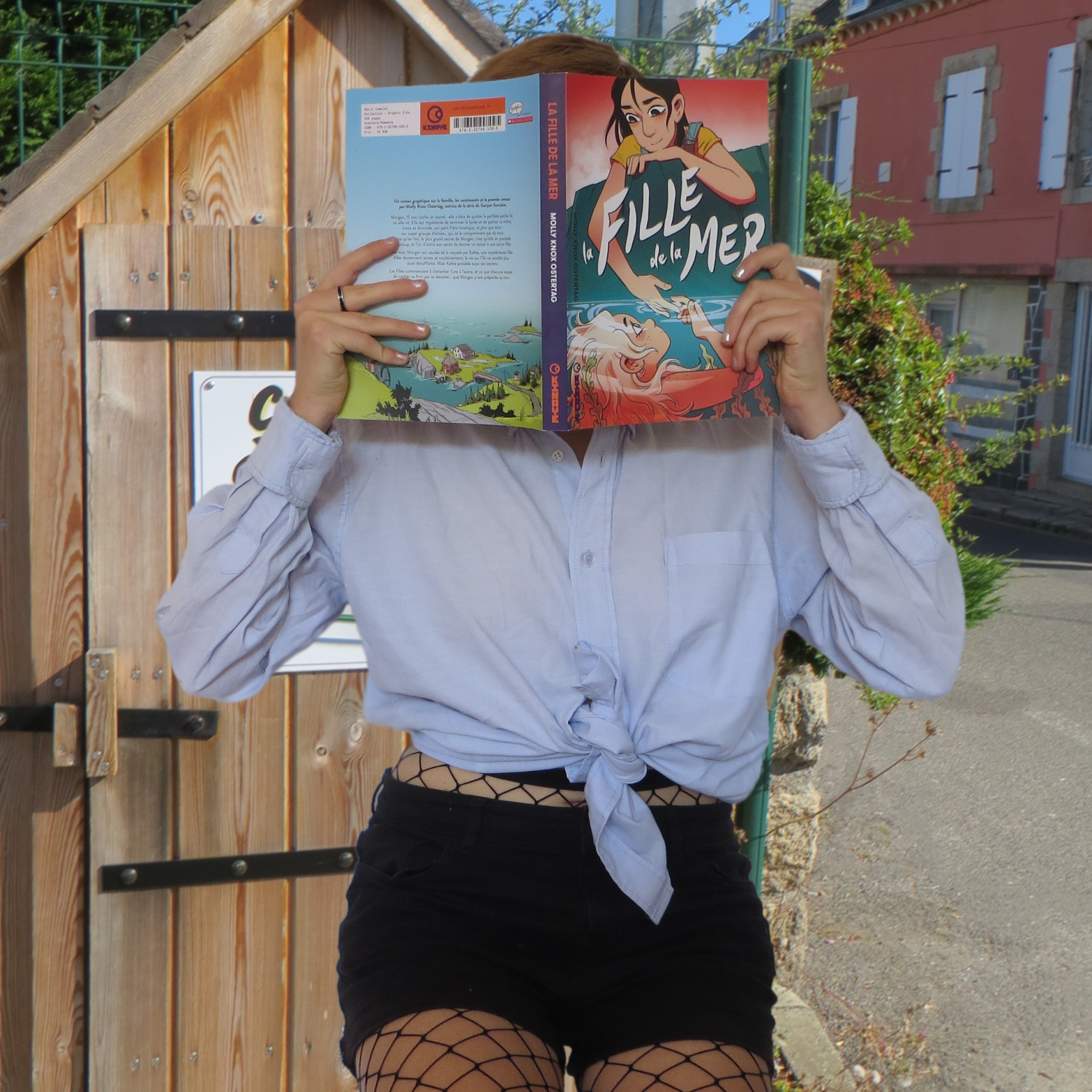 personne en chemise et collant à motif filet lisant La Fille de la Mer de Molly Knox Ostertag devant une cabane à livres en bord de mer