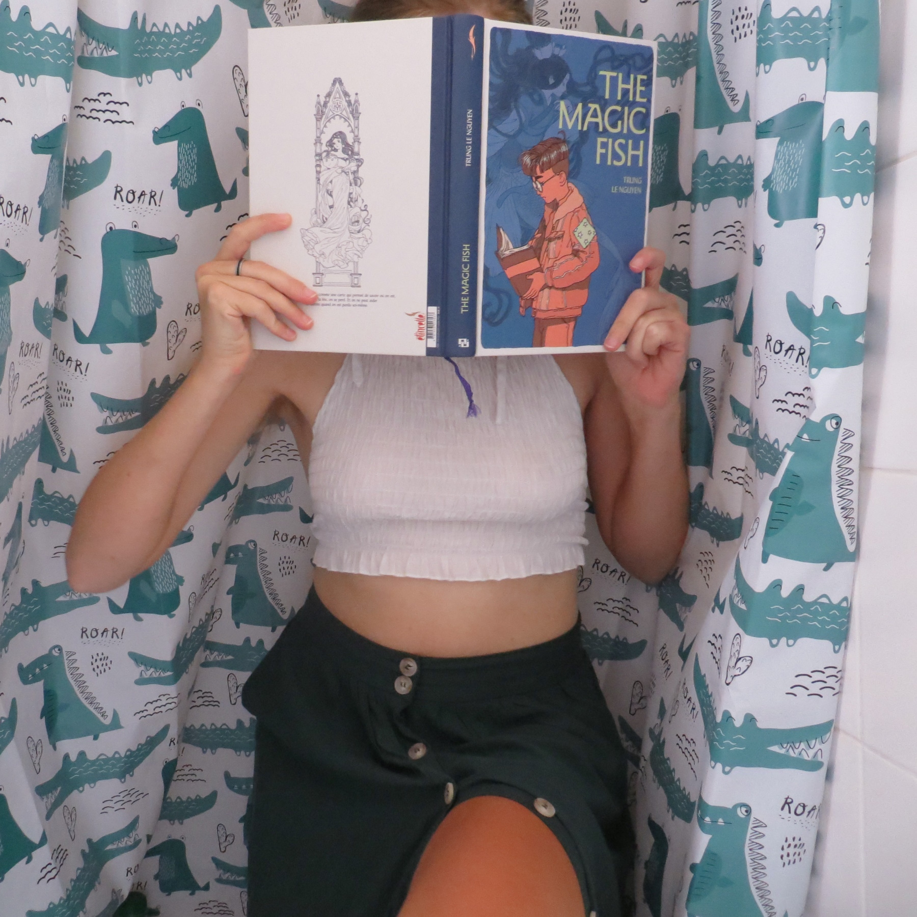 personne en crop top et longue jupe verte lisant The Magic Fish de Trung Le Nguyen dans une douche avec un rideau décoré de crocodiles