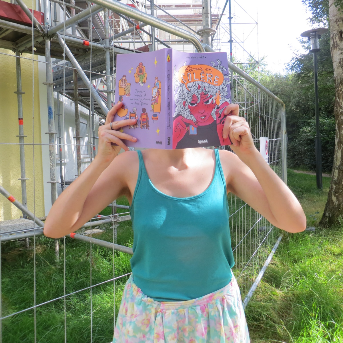personne en débardeur et short à fleurs lisant Clémence en Colère de Mirion Malle devant des grilles et échafaudages de chantier