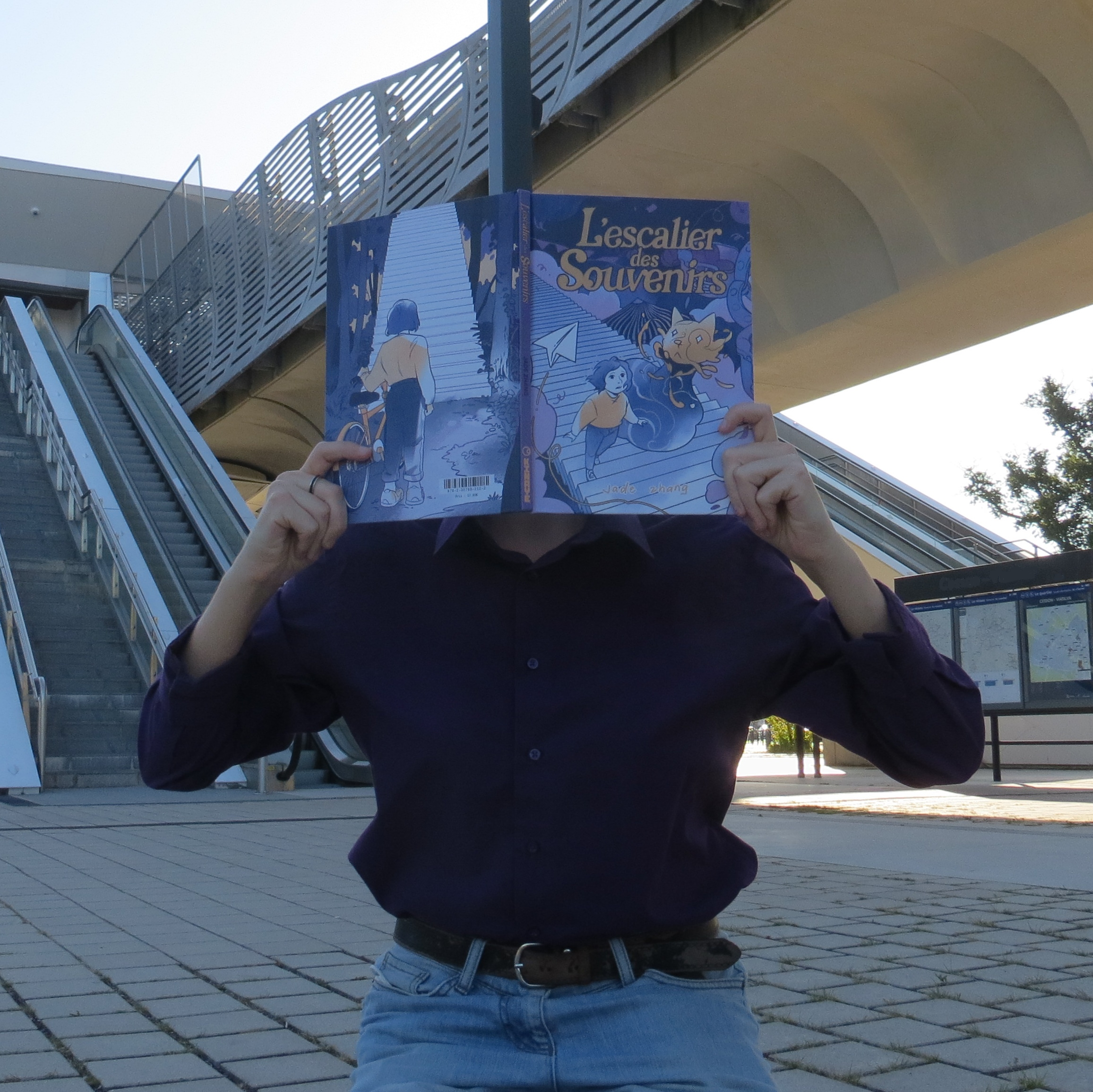 personne en chemise violette lisant L'escalier des Souvenirs de Jade Zhang devant de multiples escalators extérieurs