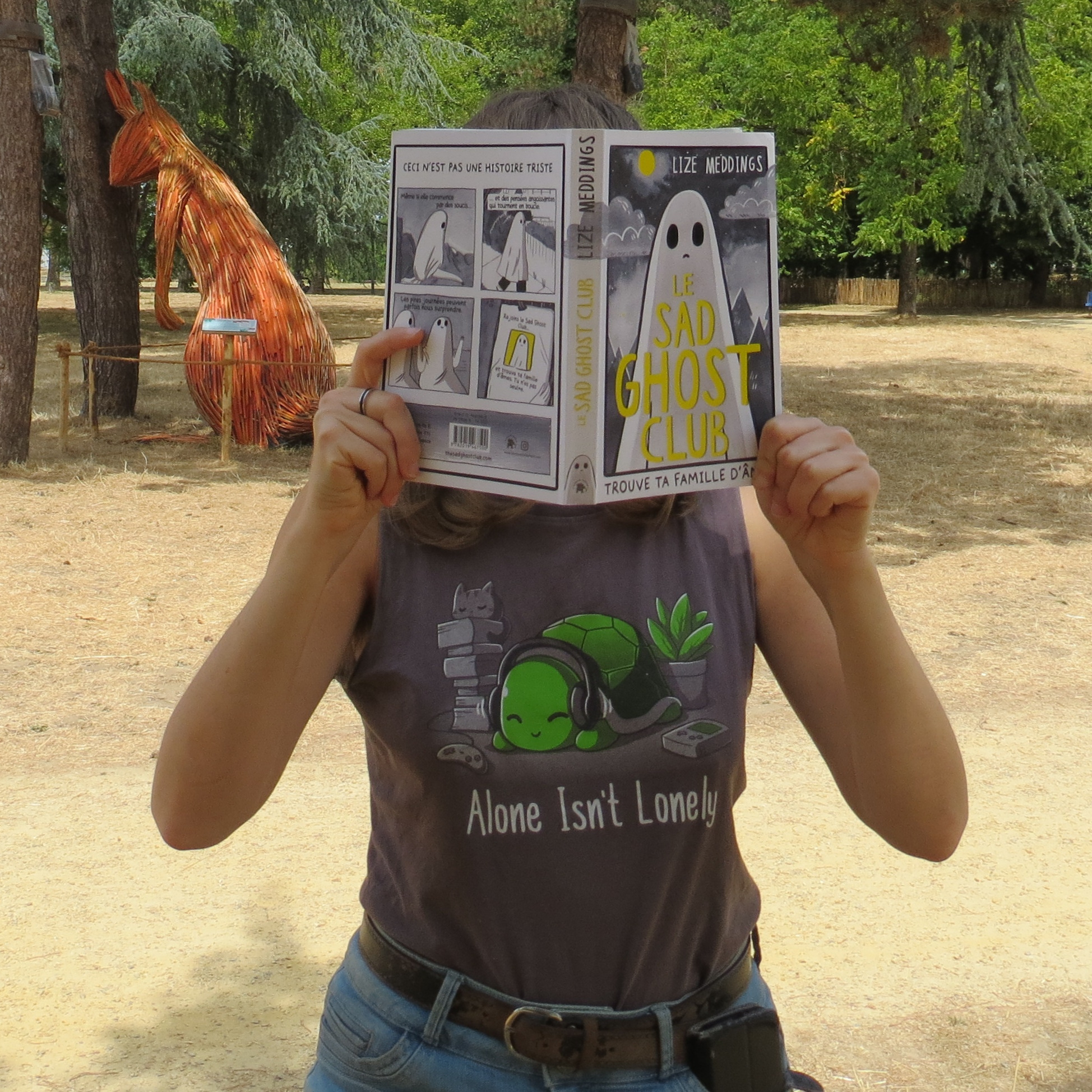 personne en débardeur avec une tortue et le sous-titre alone isn't lonely lisant Sad Ghost Club tome 1 de Lize Meddings devant un parc avec un écureuil géant s'appuyant d'un air fatigué contre les arbres