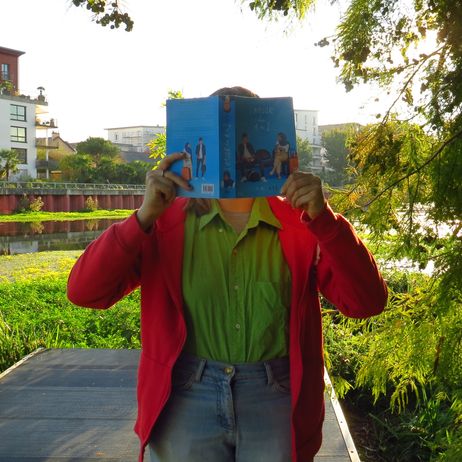 personne en chemise verte et gilet rouge lisant L’amour de A à Z de S.K. Ali devant la confluence de deux cours d'eau