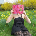 personne en robe noire et collants à dentelle lisant The Black Flamingo de Dean Atta au milieu de fleurs jaunes
