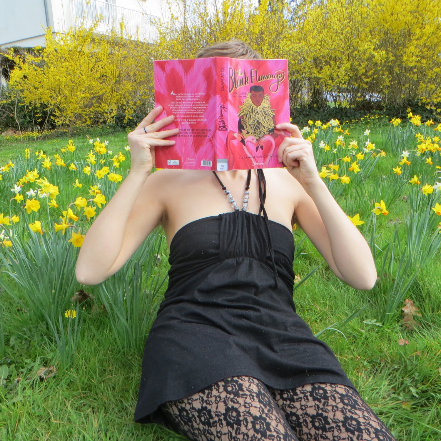 personne en robe noire et collants à dentelle lisant The Black Flamingo de Dean Atta au milieu de fleurs jaunes