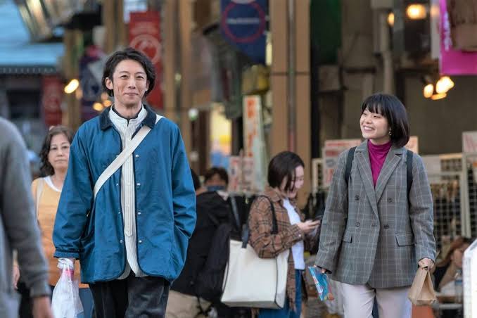 les deux personnages principaux de Koisenu Futari marchant dans la rue, avec un large espace entre eux et une expression réjouie